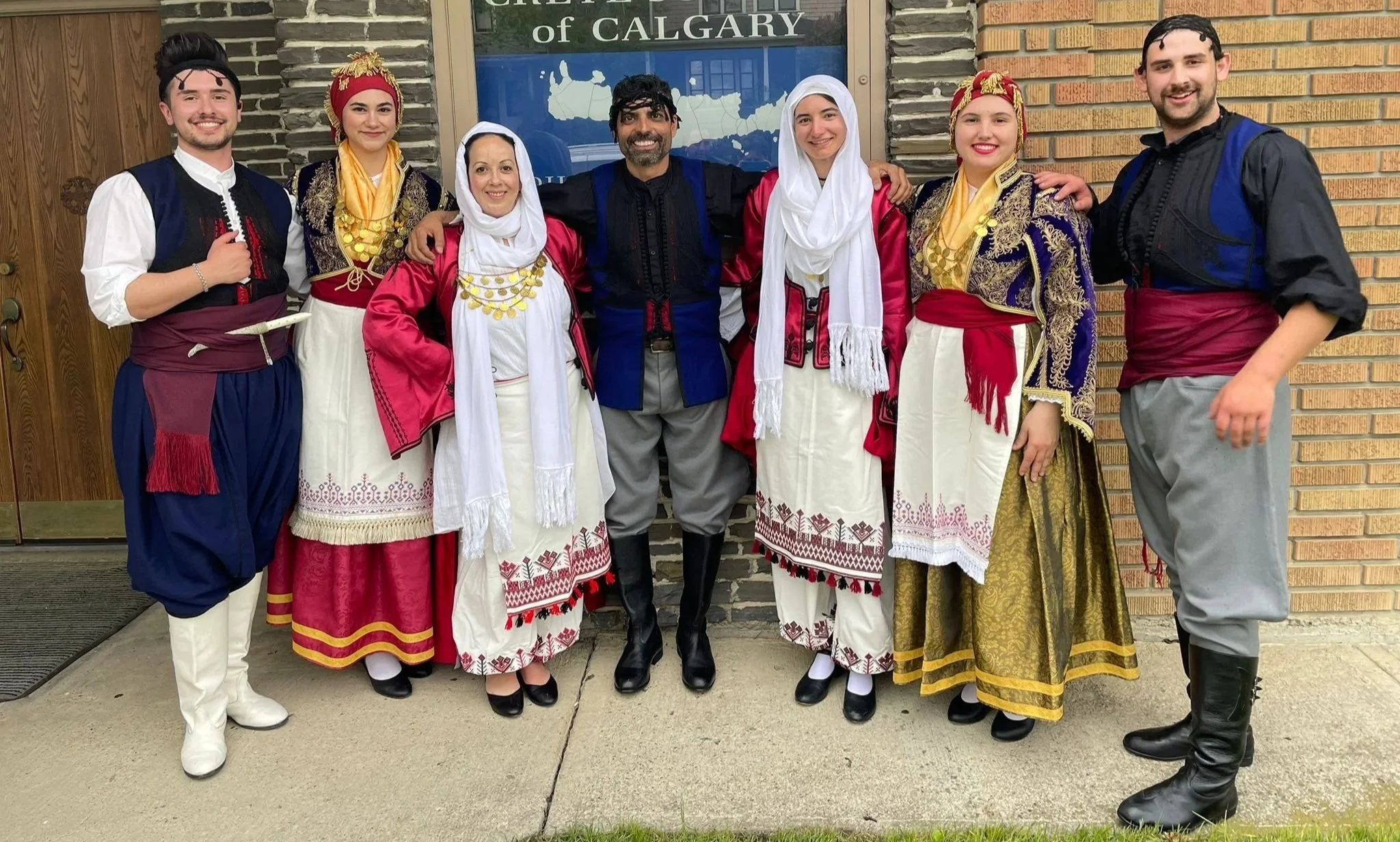 Group of eight people dressed in traditional clothing standing in front of a building with a sign that reads 'Chamber of Calgary'.