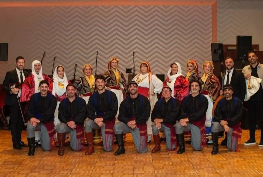 Group of people in traditional Greek costumes posing for a photo in a hall with a chevron wall and wooden floor.