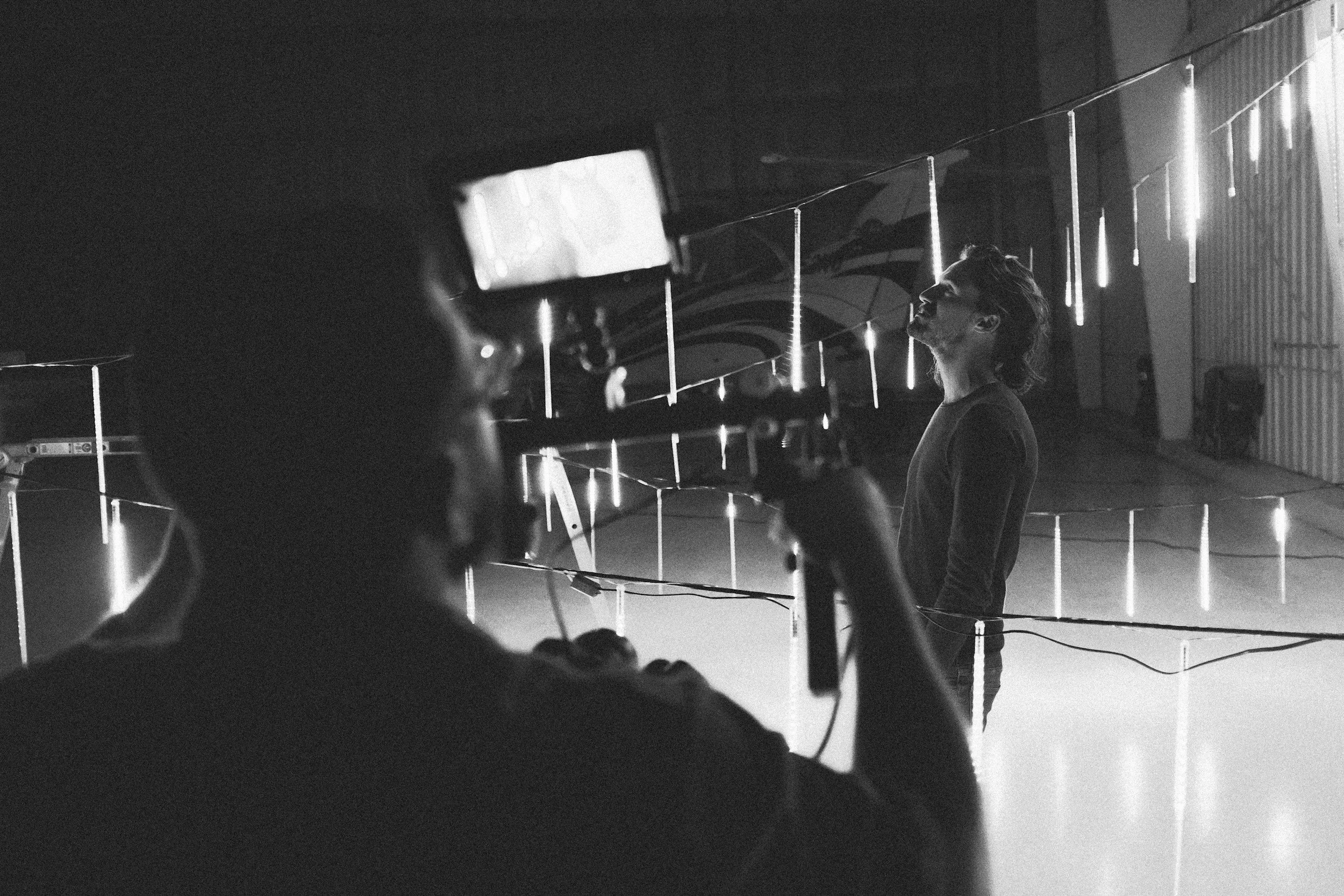 A woman with curly hair is standing in front of a wall with hanging lights, possibly during a photoshoot or film set, with a person in the foreground operating filming equipment.
