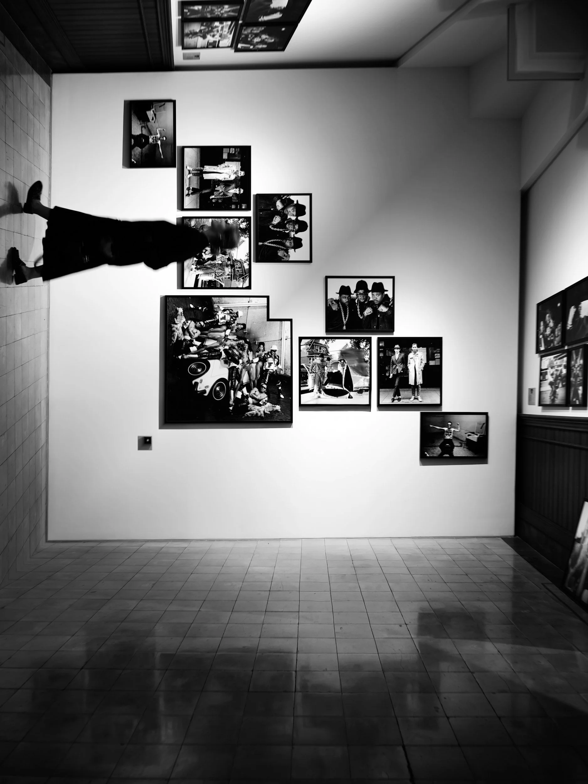 Black and white photograph of a person walking past an art gallery wall displaying multiple framed photographs.
