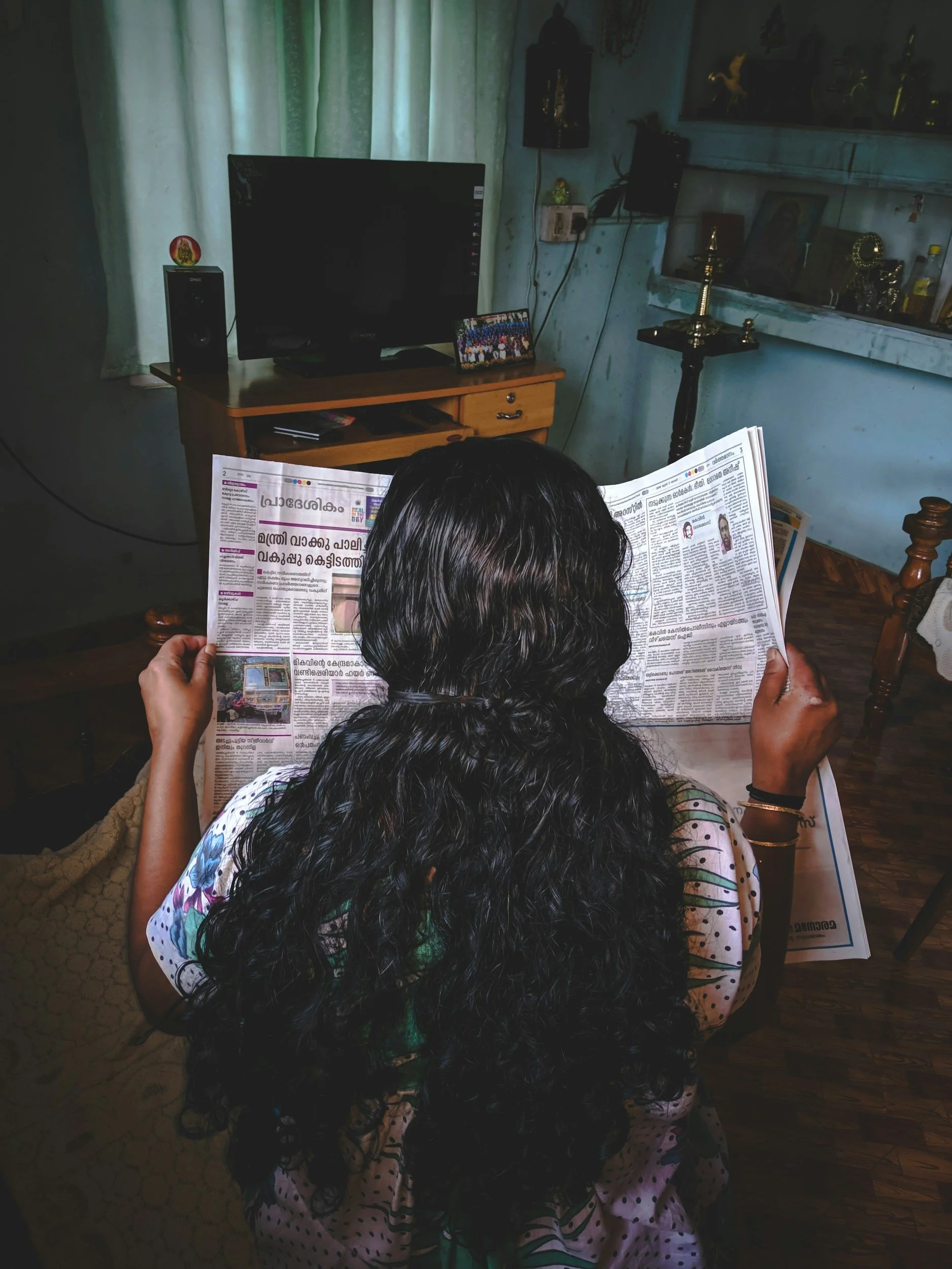 A woman with long dark hair sitting in a room, reading a newspaper, with a TV and other items in the background.