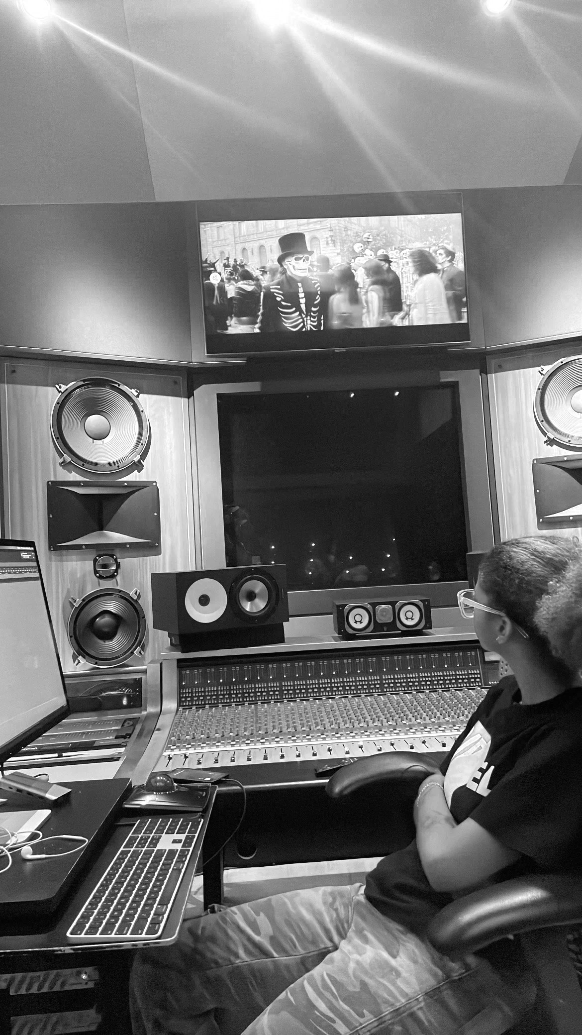A person sitting at a sound mixing console in a recording or studio environment, wearing glasses and a dark shirt, with studio speakers, a computer, and audio equipment visible around them. A TV screen above shows a black and white image of a crowd, including a person dressed as a skeleton and wearing a top hat.