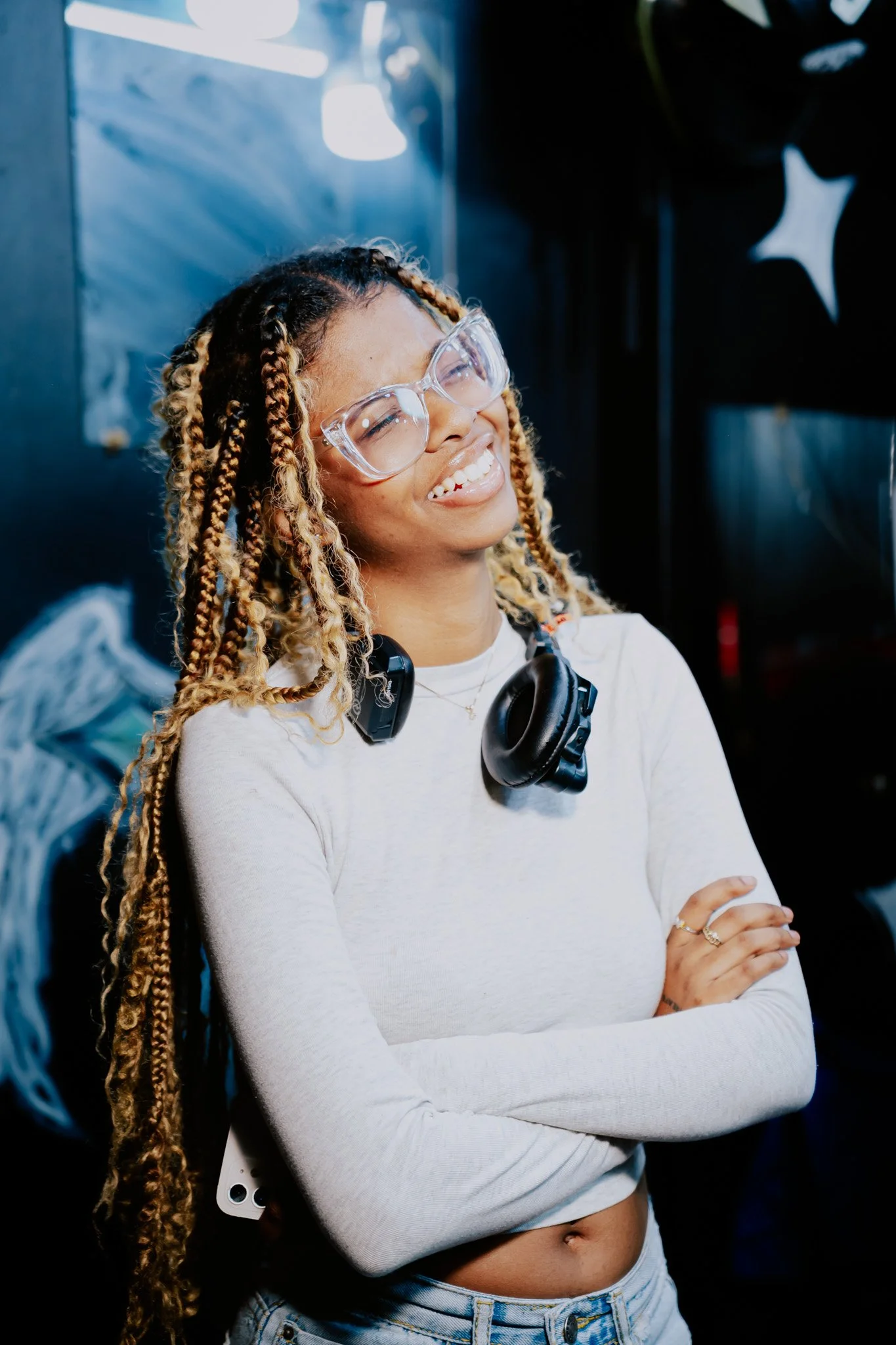 A young woman with long braids wearing large glasses, headphones around her neck, a white long-sleeve top, and light blue jeans. She is smiling with her eyes closed, arms crossed, standing against a dark background.