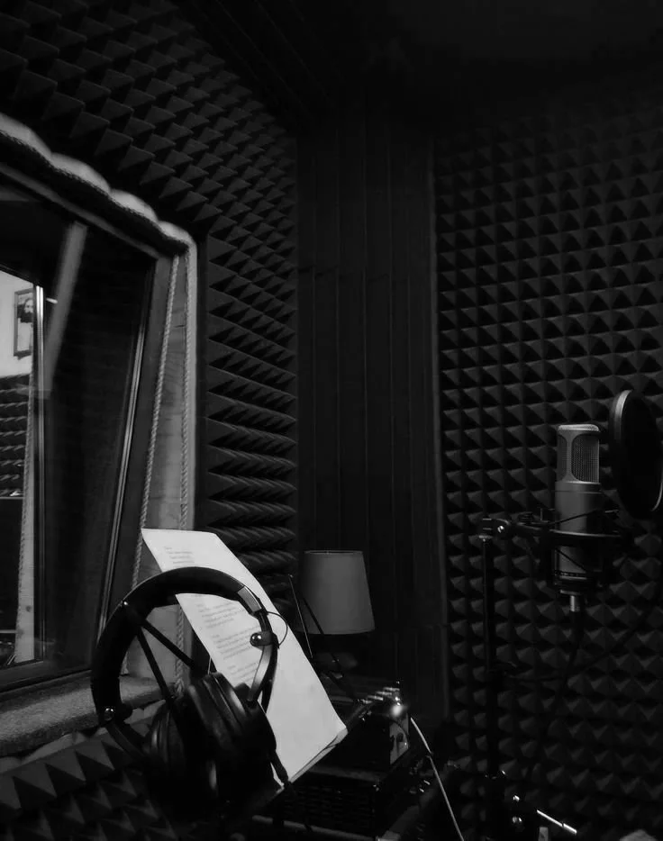 Recording studio with black acoustic foam walls, a microphone with pop filter, headphones on a stand, a music stand with sheet music, and a window with soundproofing.