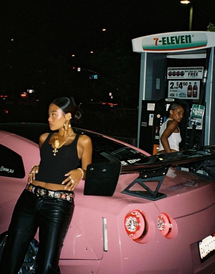 Two women at a 7-Eleven gas station at night, with a pink sports car featuring a large rear spoiler in the foreground.