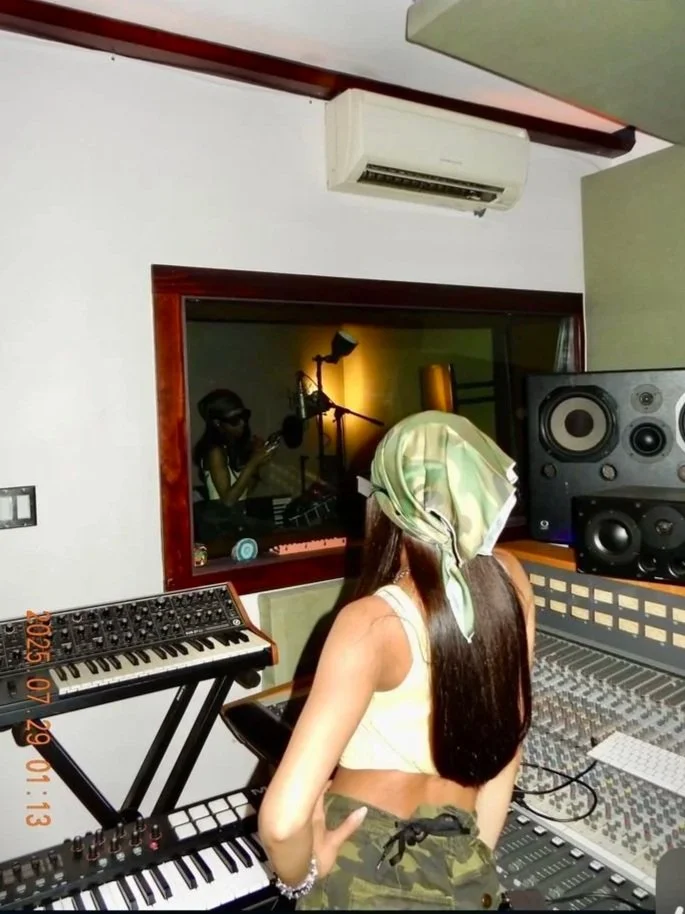 A woman with long brown hair, wearing a camouflage headscarf and camouflage shorts, is working in a recording studio surrounded by music equipment, including a keyboard, mixing console, and speakers. There is a woman singing in a booth visible through a glass window, with warm lighting.