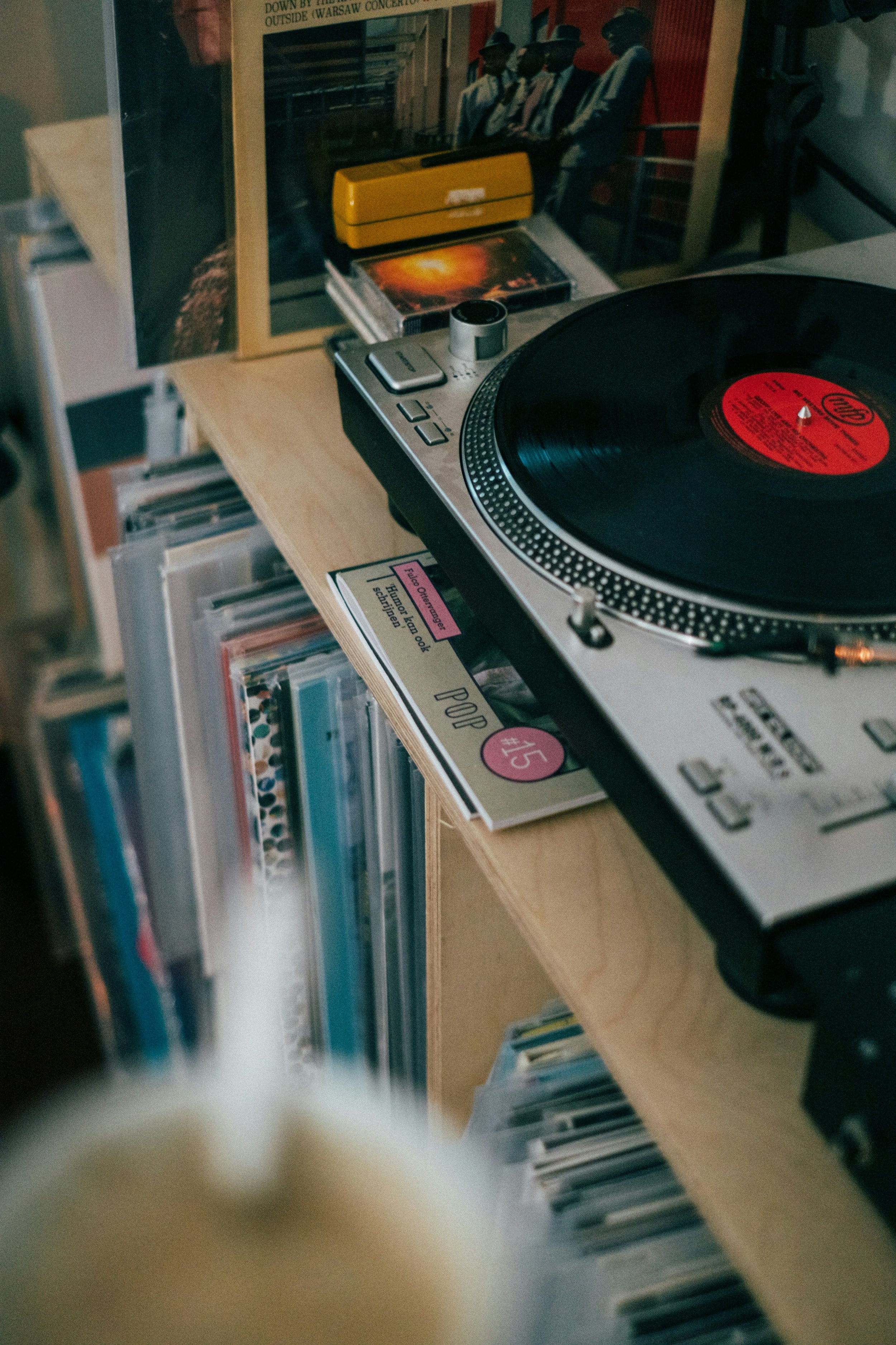 A turntable playing a vinyl record on a wooden shelf with various books and magazines, including a yellow box and a film-related magazine.
