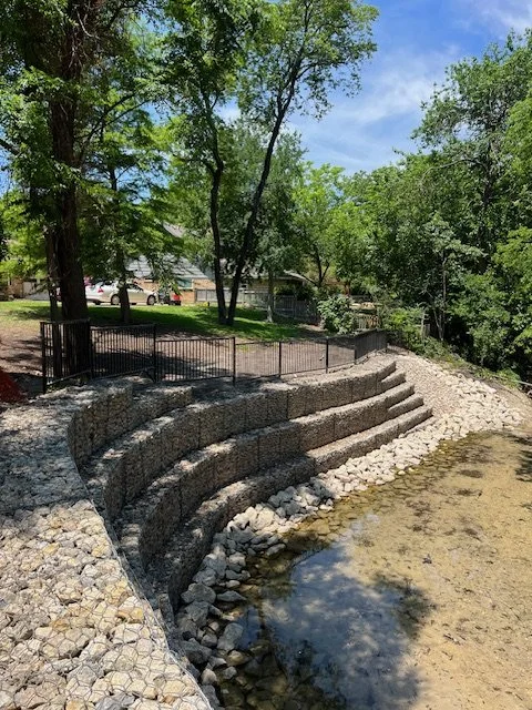 “Expert custom metal fencing installation on tiered retaining walls and erosion control systems, built for safety, strength, and long-term durability. This project showcases black steel fencing installed along stepped retaining walls near a creek, pr