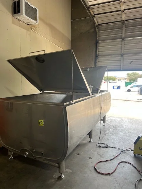 A large stainless steel dairy tank that has been converted for industrial use, featuring hinged lids and a polished finish. Located inside a workshop, the tank is designed for bulk liquid processing.