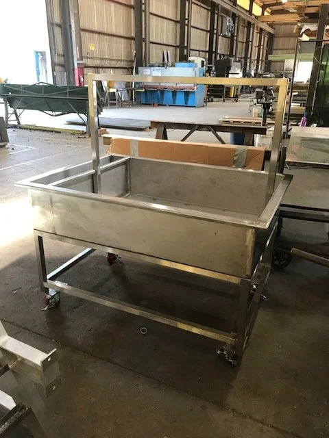 A custom stainless steel dual compartment tank mounted on a heavy-duty wheeled frame inside a fabrication shop. Designed for mobility and efficiency, the unit features a durable structure suitable for industrial washing, processing, or liquid handlin