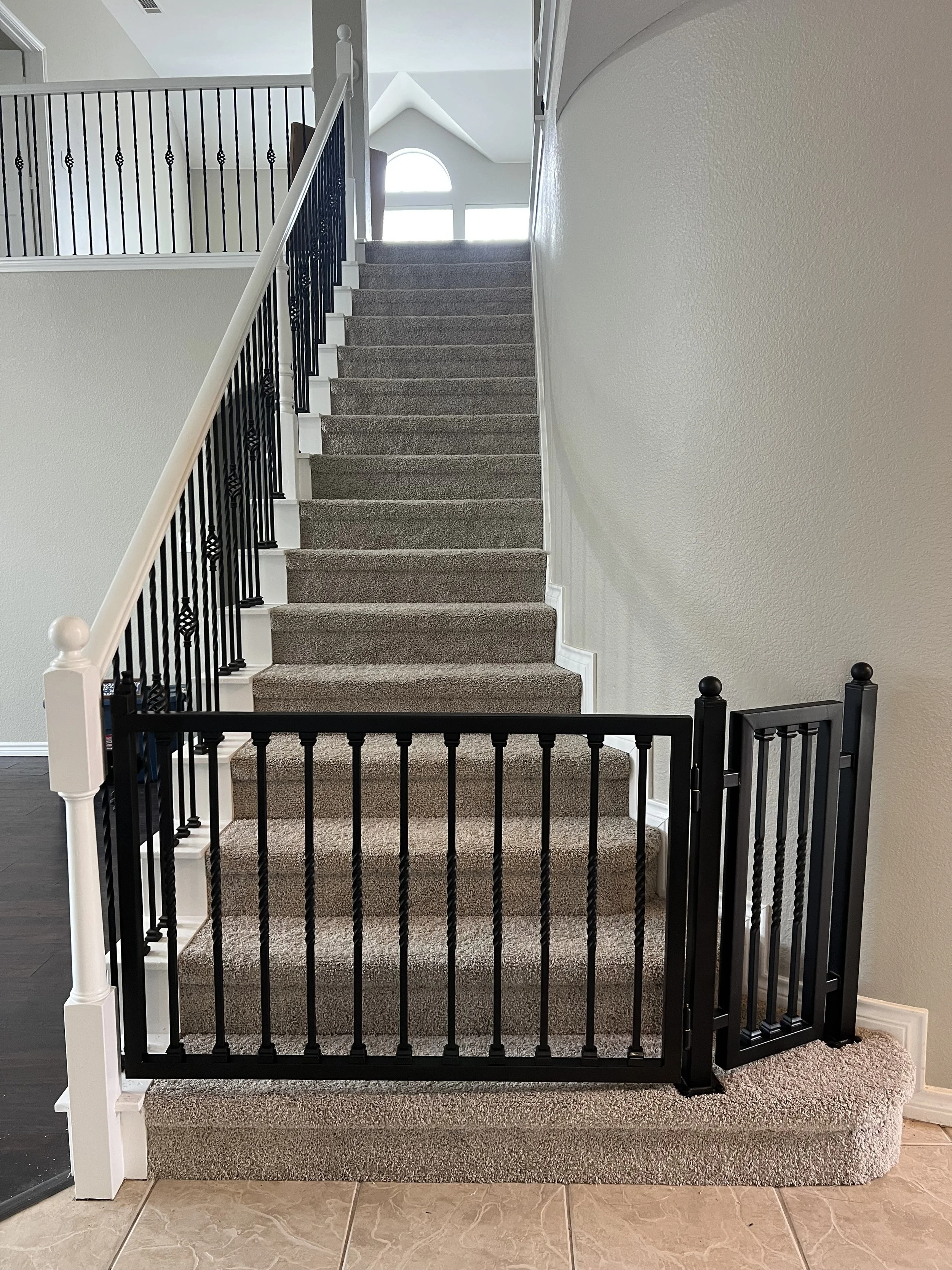 Custom-installed black metal child safety gate mounted at the base of a carpeted staircase, featuring secure hardware-mounted posts, durable vertical balusters, and a convenient walk-through door. Designed to blend with modern home interiors, this st