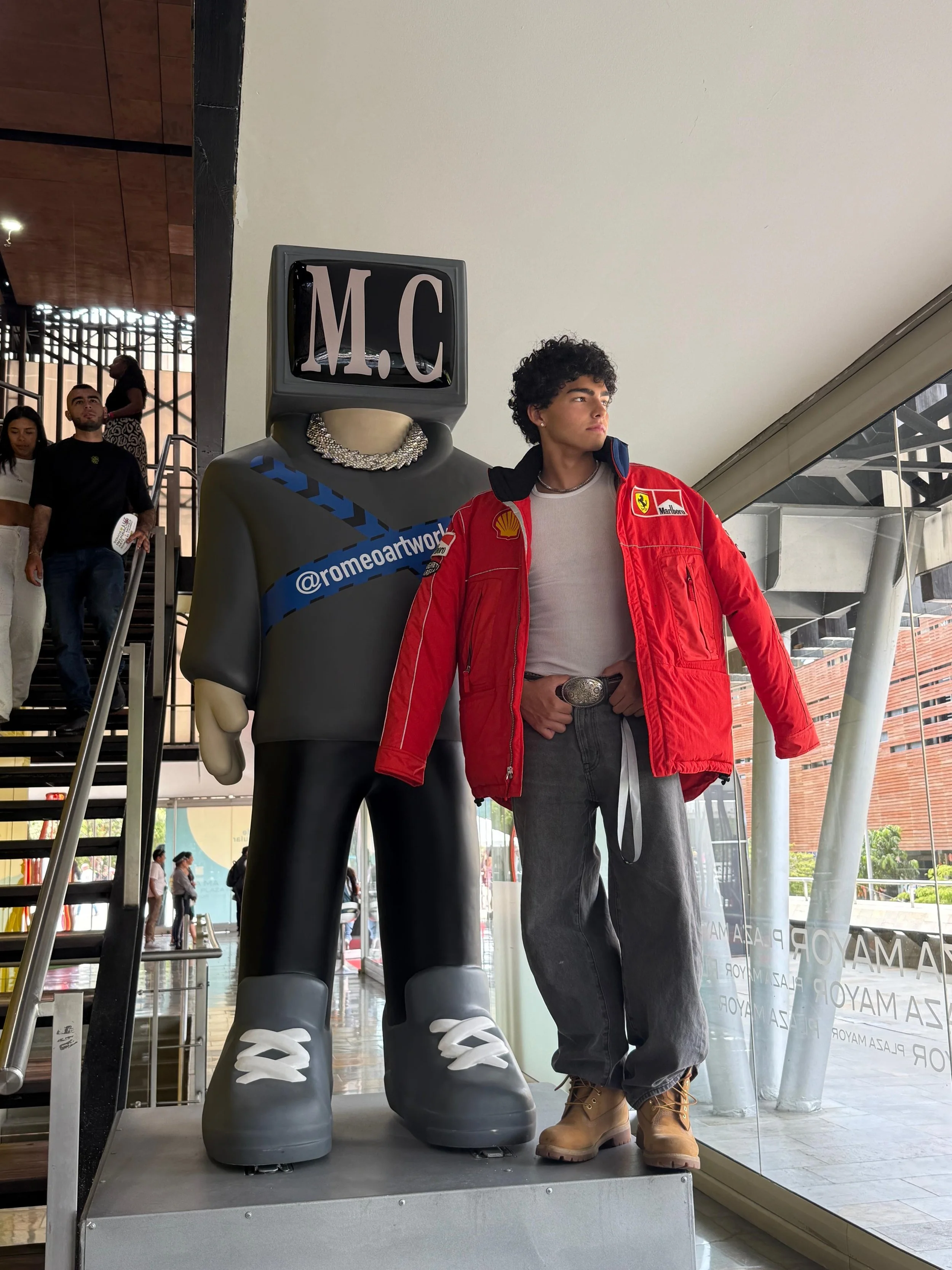 A young man posing next to a large humanoid statue with a television for a head displaying the initials 'M.C'. The statue is dressed stylishly with a shiny necklace and gray shoes, and the young man is wearing a red Ferrari jacket, gray pants, and tan boots.