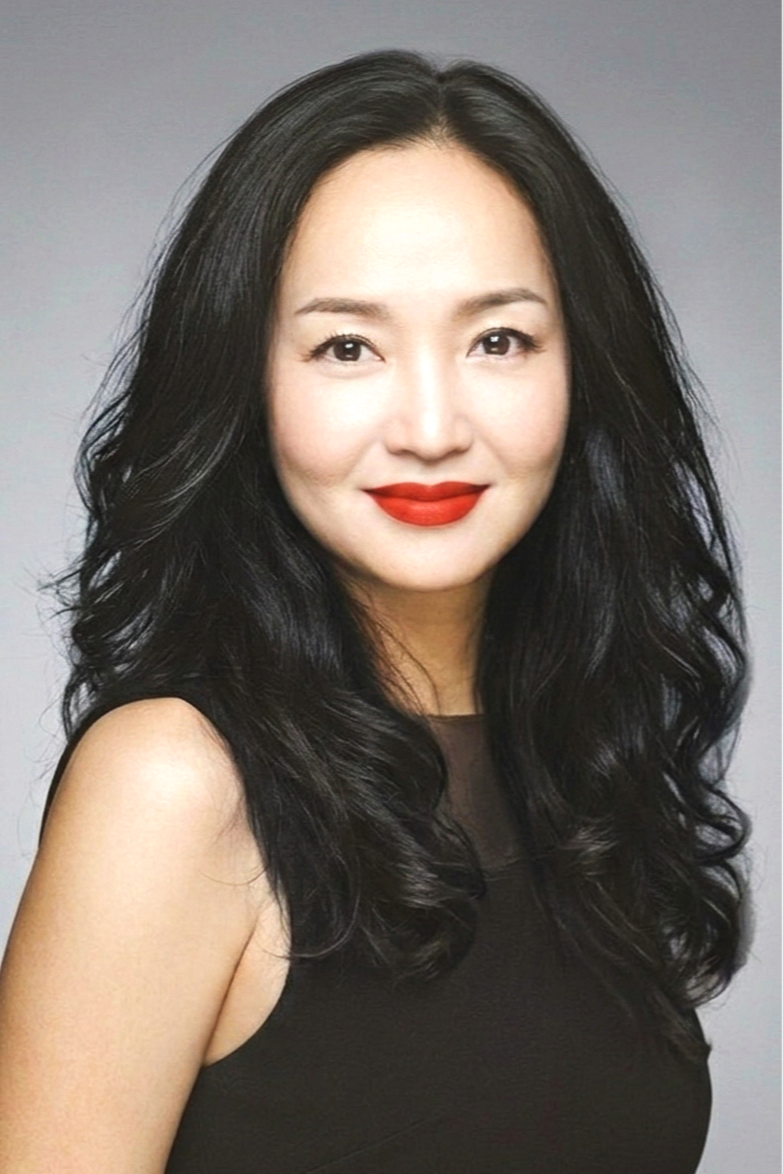 A portrait of a woman with long black curly hair, red lipstick, and a black sleeveless top, smiling at the camera against a plain gray background.