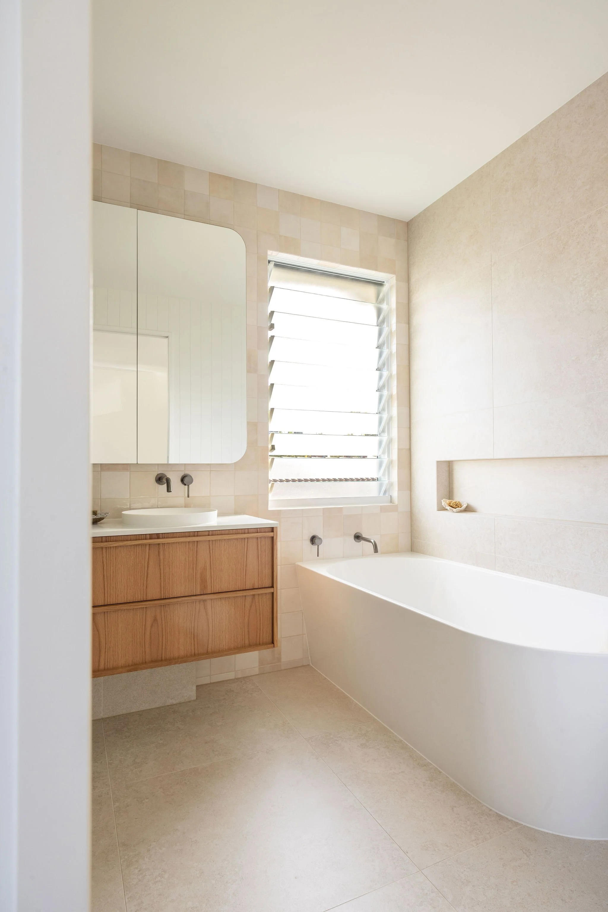 A modern bathroom with a white bathtub, wooden vanity with a vessel sink, large window with louvered blinds, and a mirror above the sink, all in neutral tones.