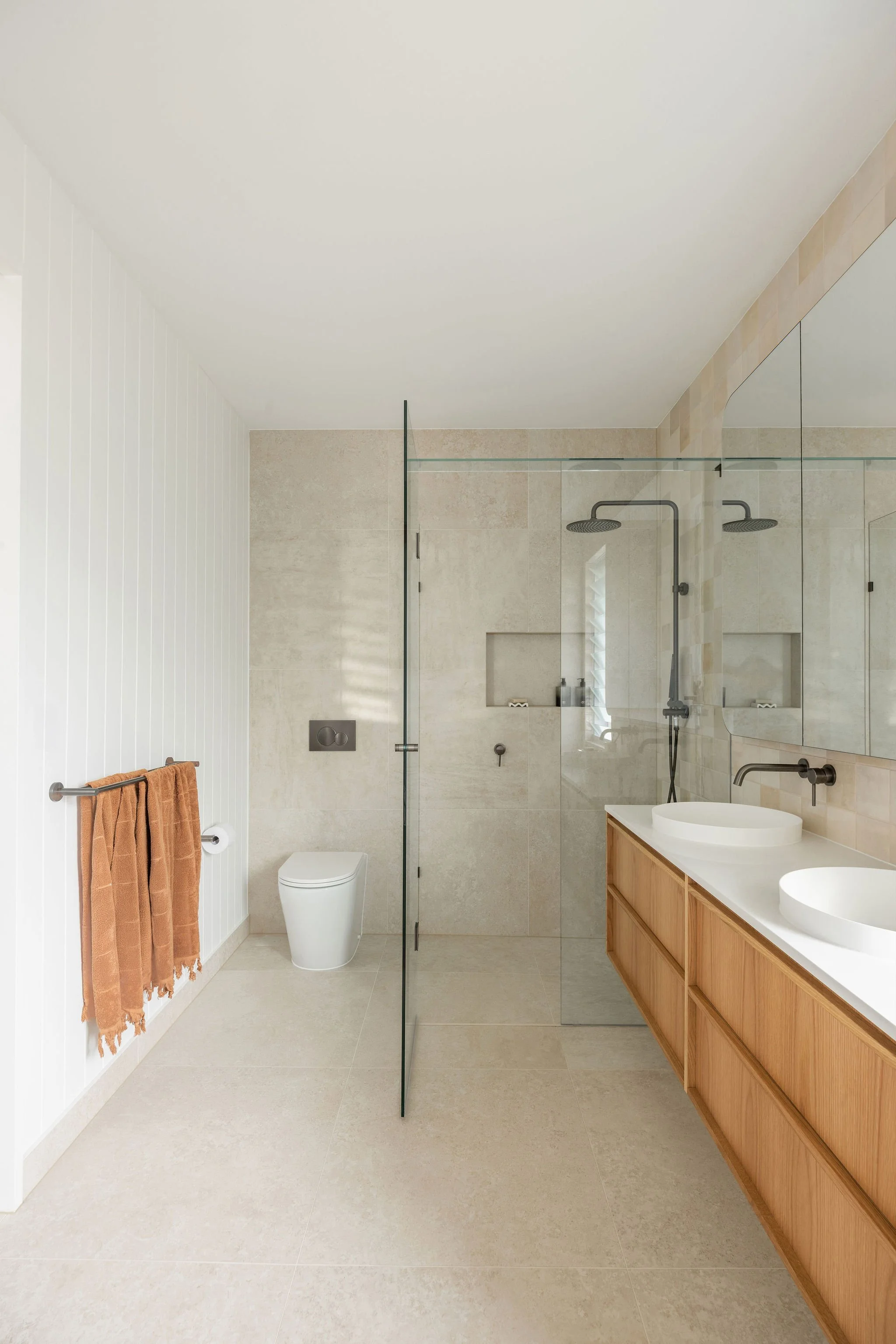 Modern bathroom with beige tile floors and walls, a glass shower enclosure with black fixtures, a white toilet, and wooden vanity with two vessel sinks and large mirror.