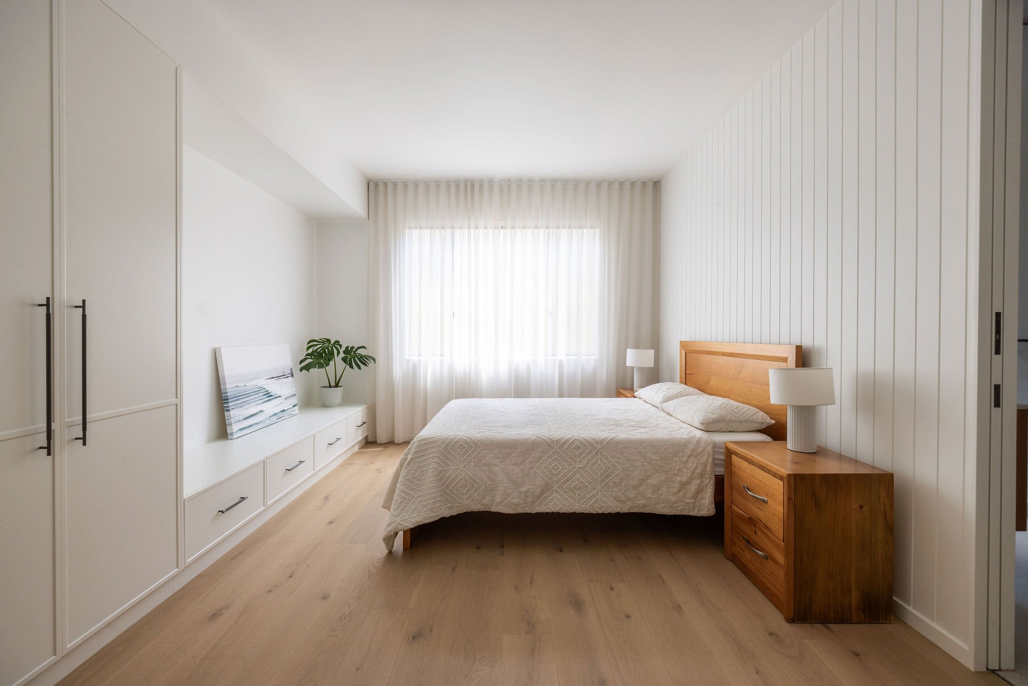 A bedroom with white walls, a large window with sheer curtains, a wooden bed with beige bedding, a wooden nightstand with a modern white lamp, and a white cabinet with drawers. There is a framed picture of a beach scene leaning against the wall and a