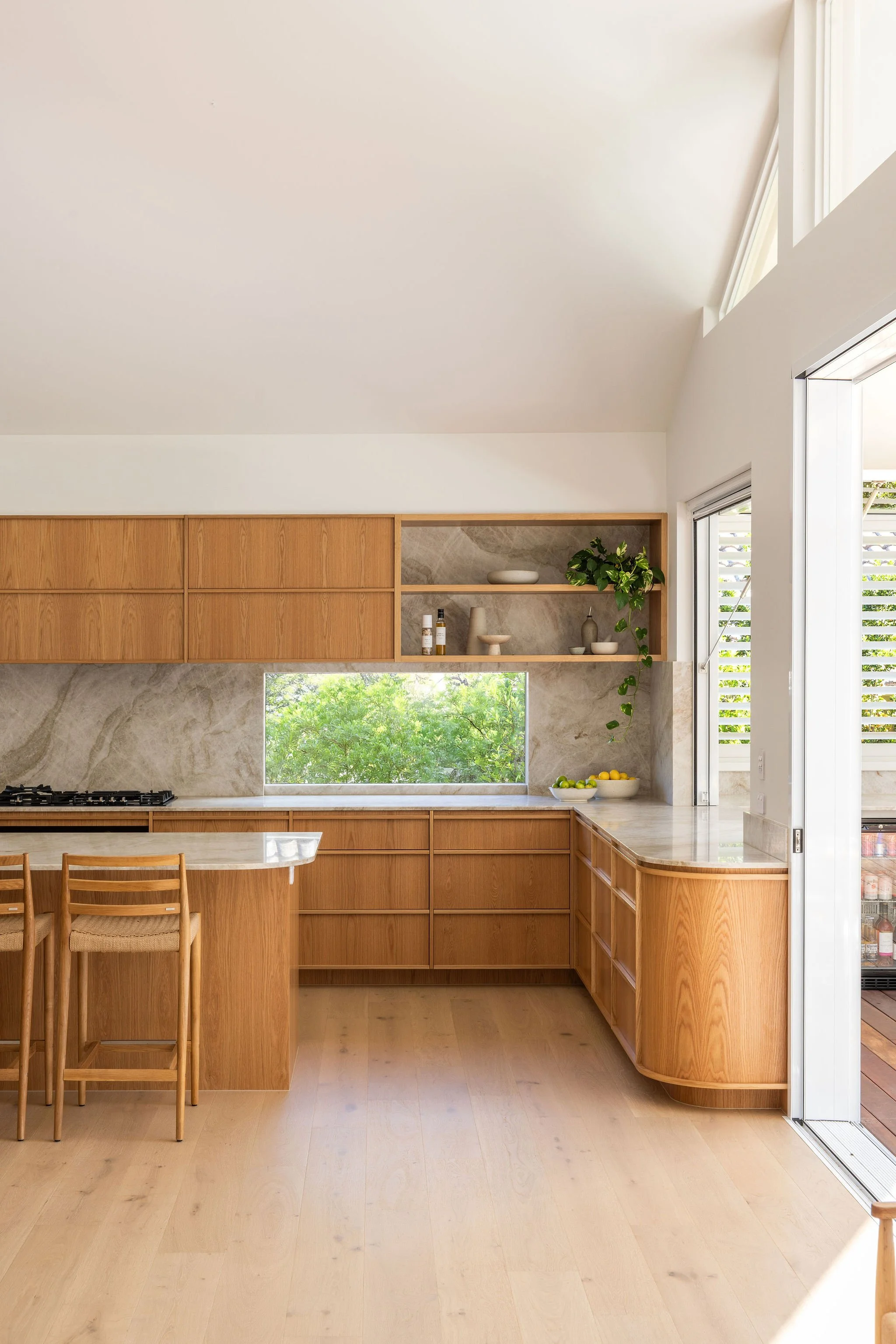 Modern kitchen with wooden cabinets, a large window overlooking greenery, and a breakfast bar with wooden chairs.