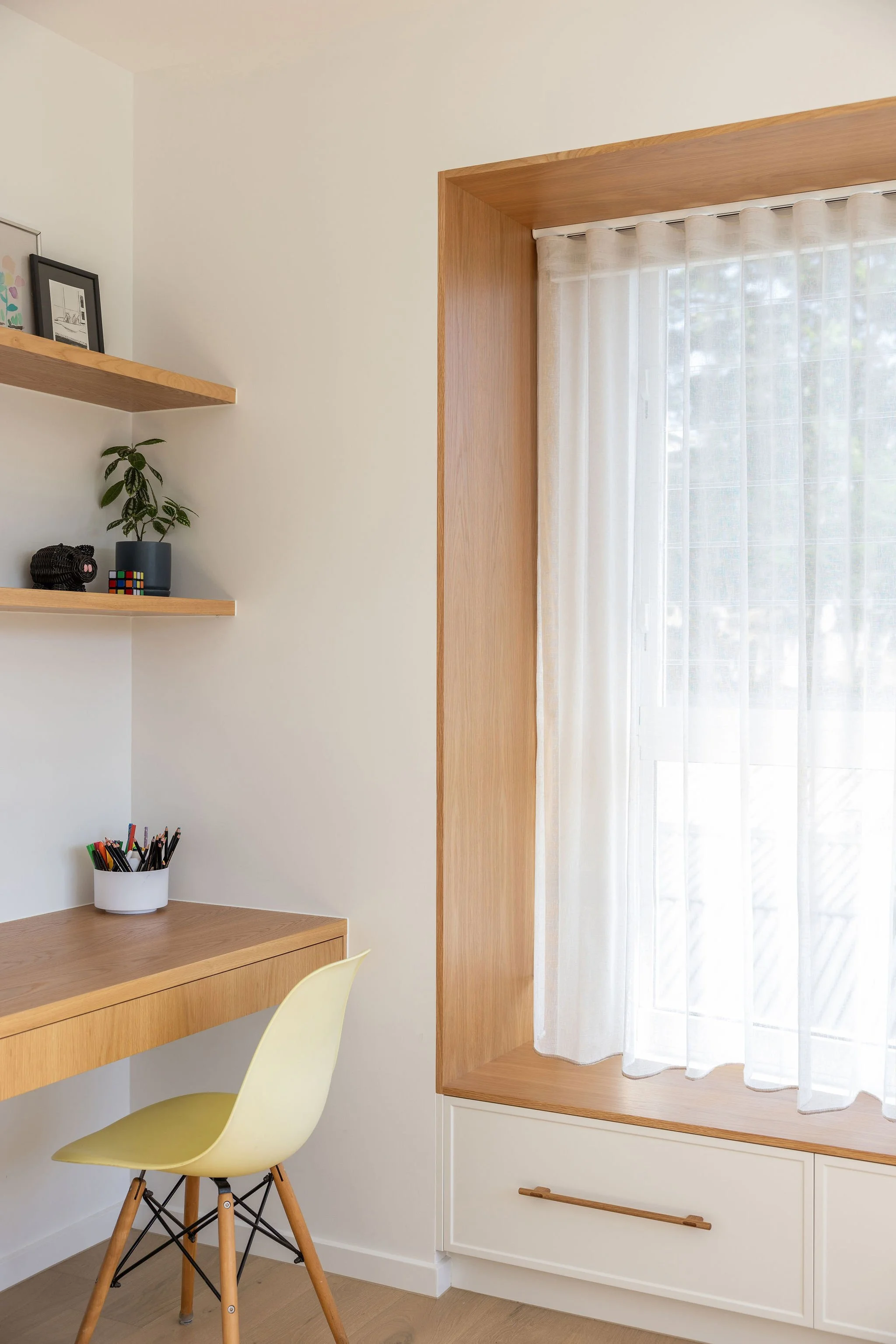 A cozy corner of a room with a wooden desk, a yellow chair, white curtains, a small floating shelf with framed pictures and a plant, and a window with natural light.