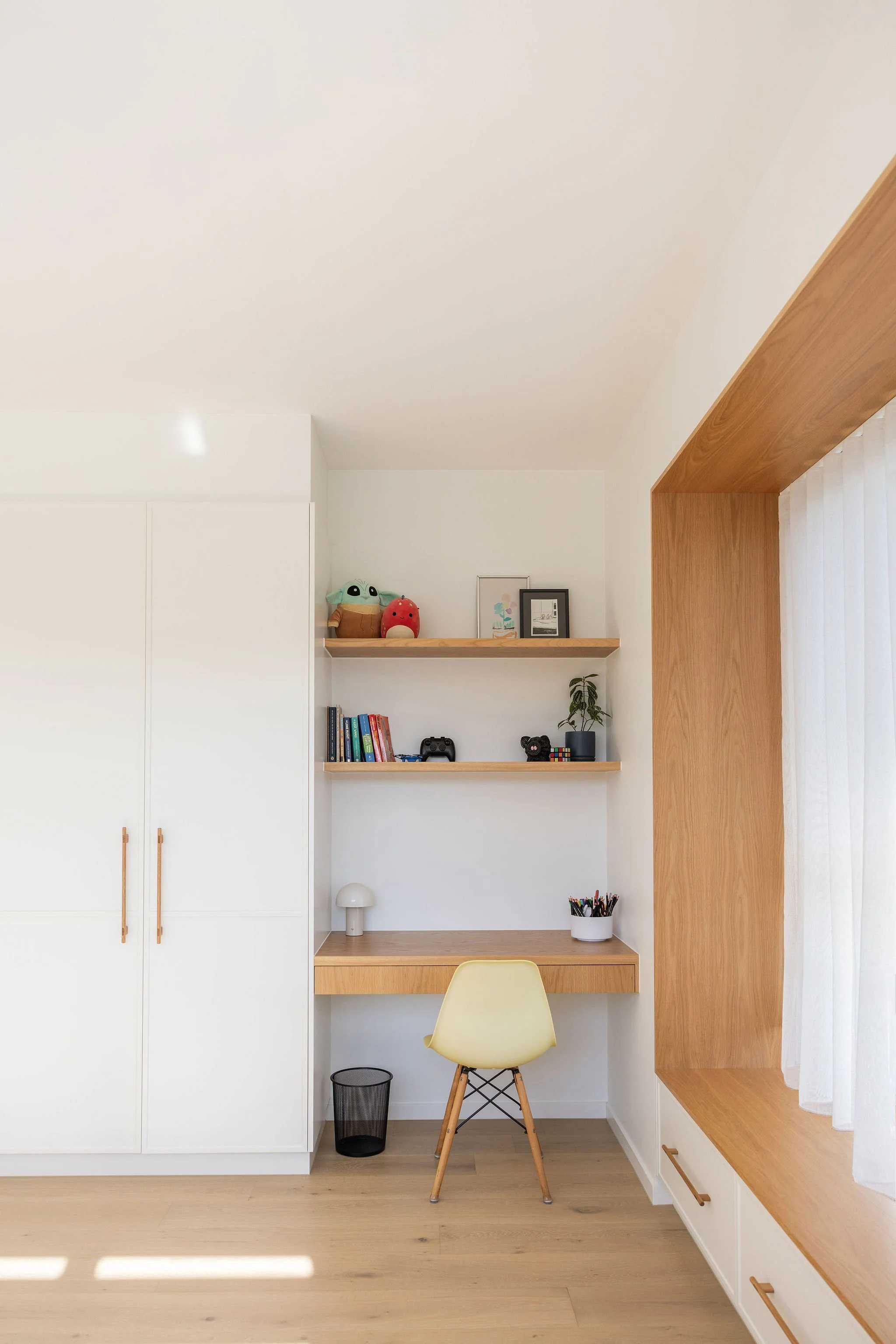 A minimalist home office nook with white cabinets and wooden shelves, decorated with books, framed art, plush toys, and a potted plant, illuminated by natural light through a large window with sheer curtains.