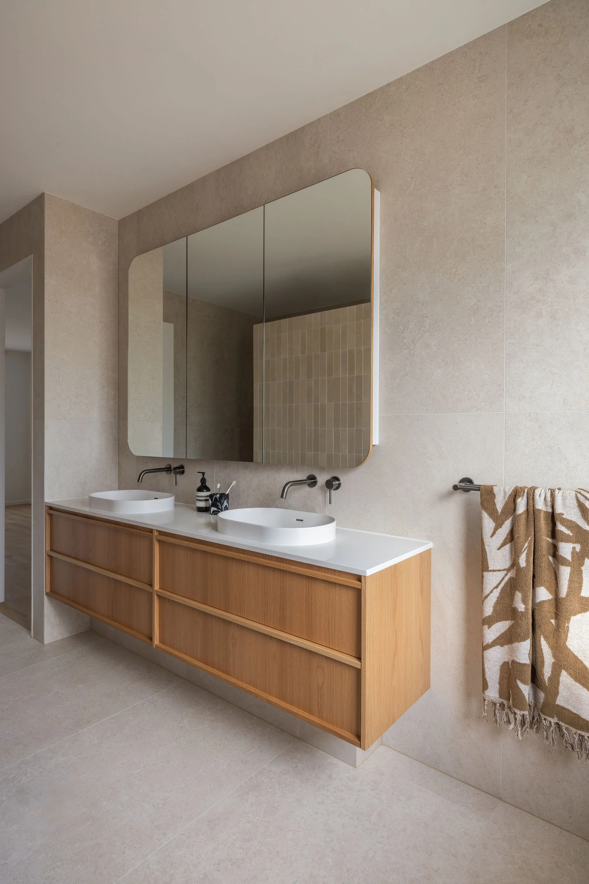 Modern bathroom with two sinks, wooden vanity, large mirror, beige tiled walls, and a towel hanging on a chrome towel bar.