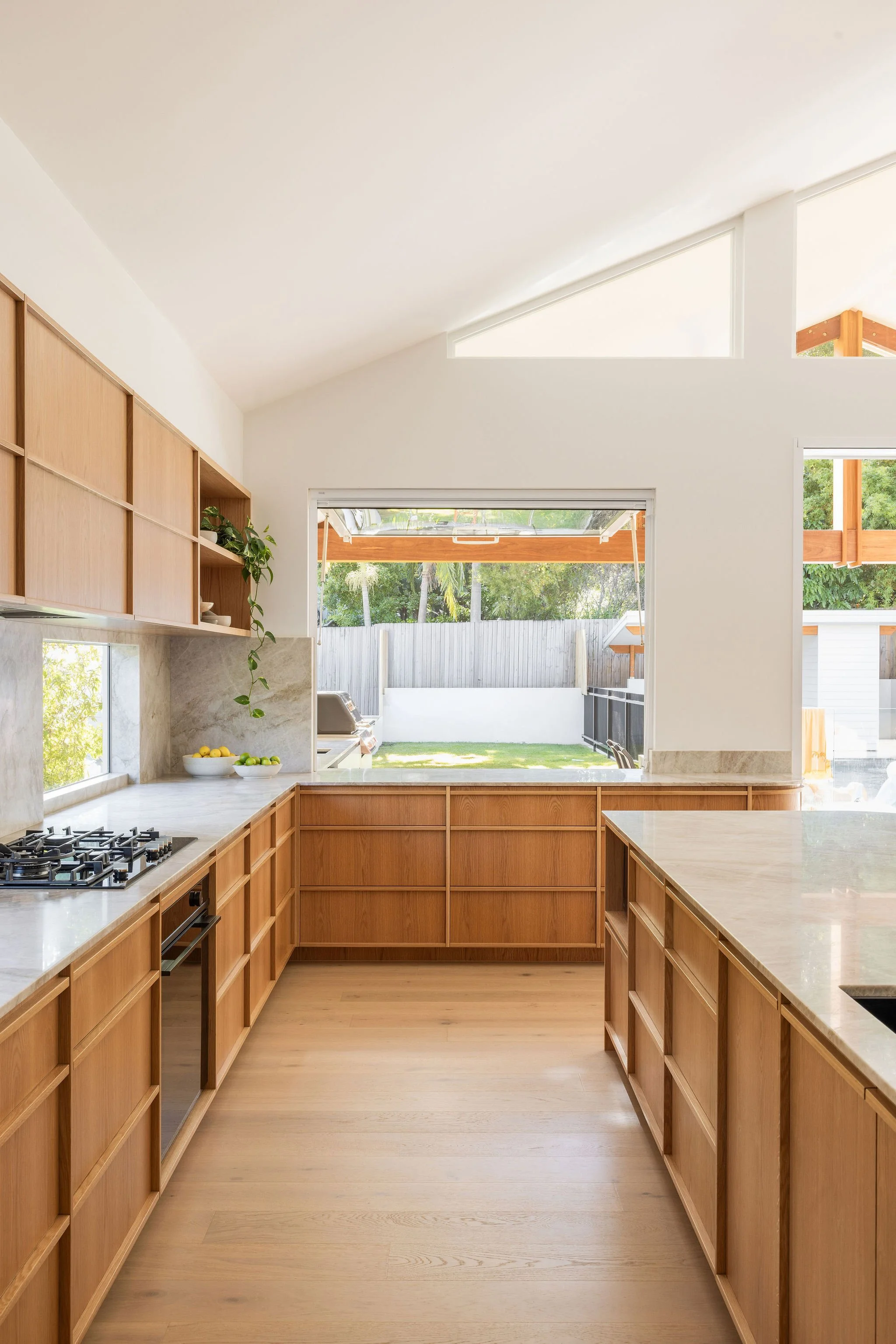 Modern kitchen with wooden cabinets, marble countertops, a large window, and a view of a backyard with a lawn and outdoor patio.
