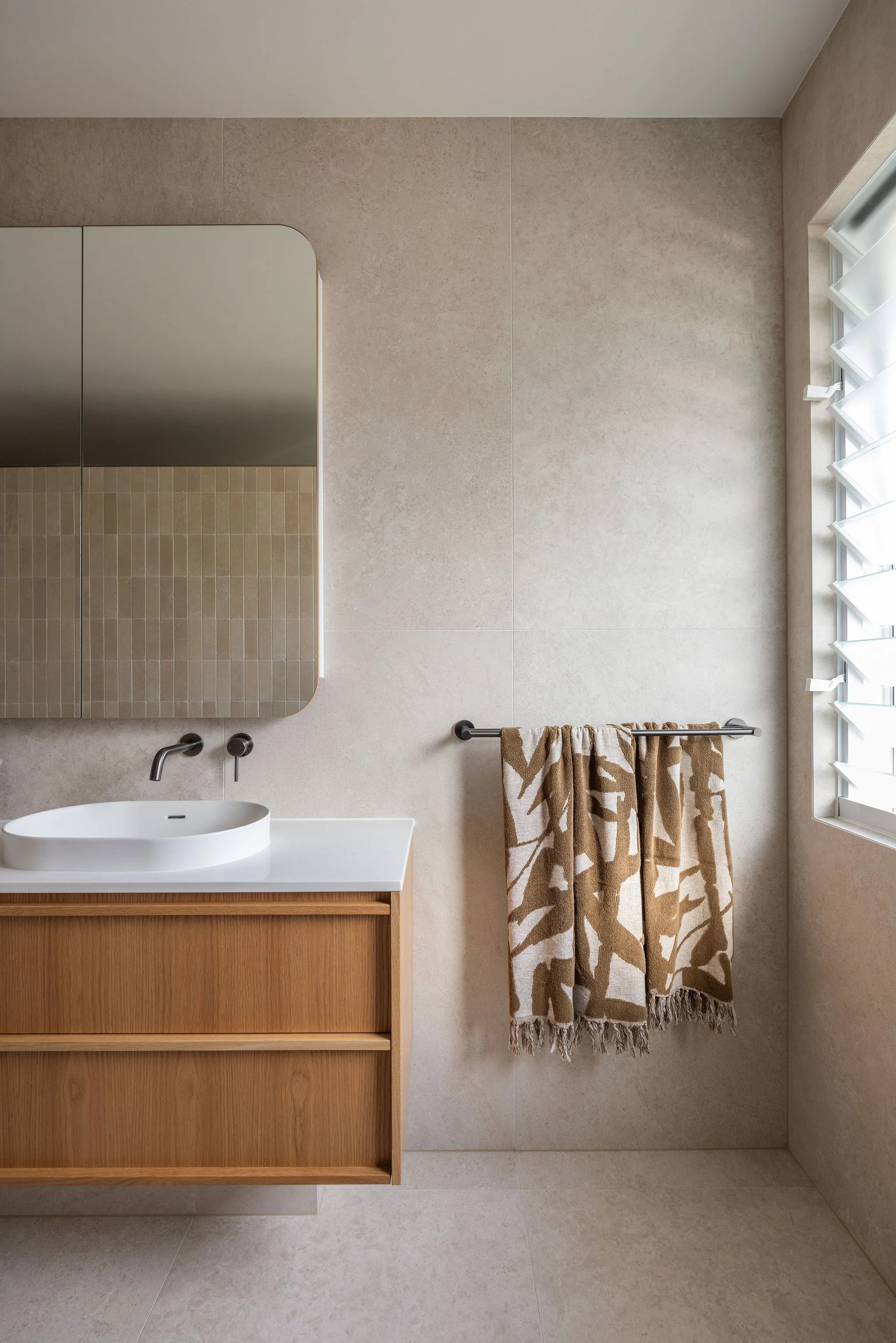 A modern bathroom with a white vessel sink on a wooden vanity, a large mirror, beige tiled walls, a window with white plantation shutters, and a beige towel with a leaf pattern hanging on a stainless steel towel bar.