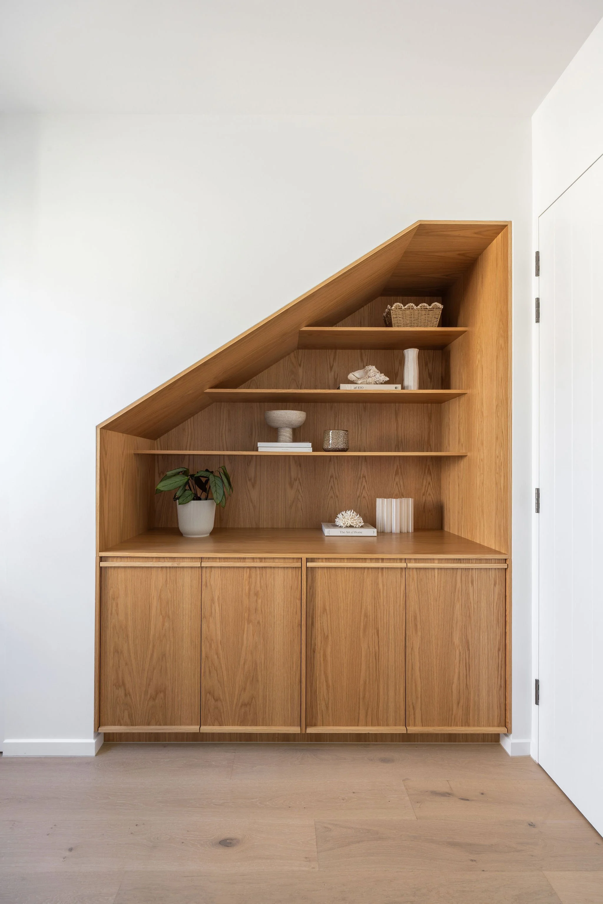 A wooden bookshelf with a sloped top and drawer at the bottom, decorated with potted plants, books, and decorative objects, against a plain white wall and light-colored floor.