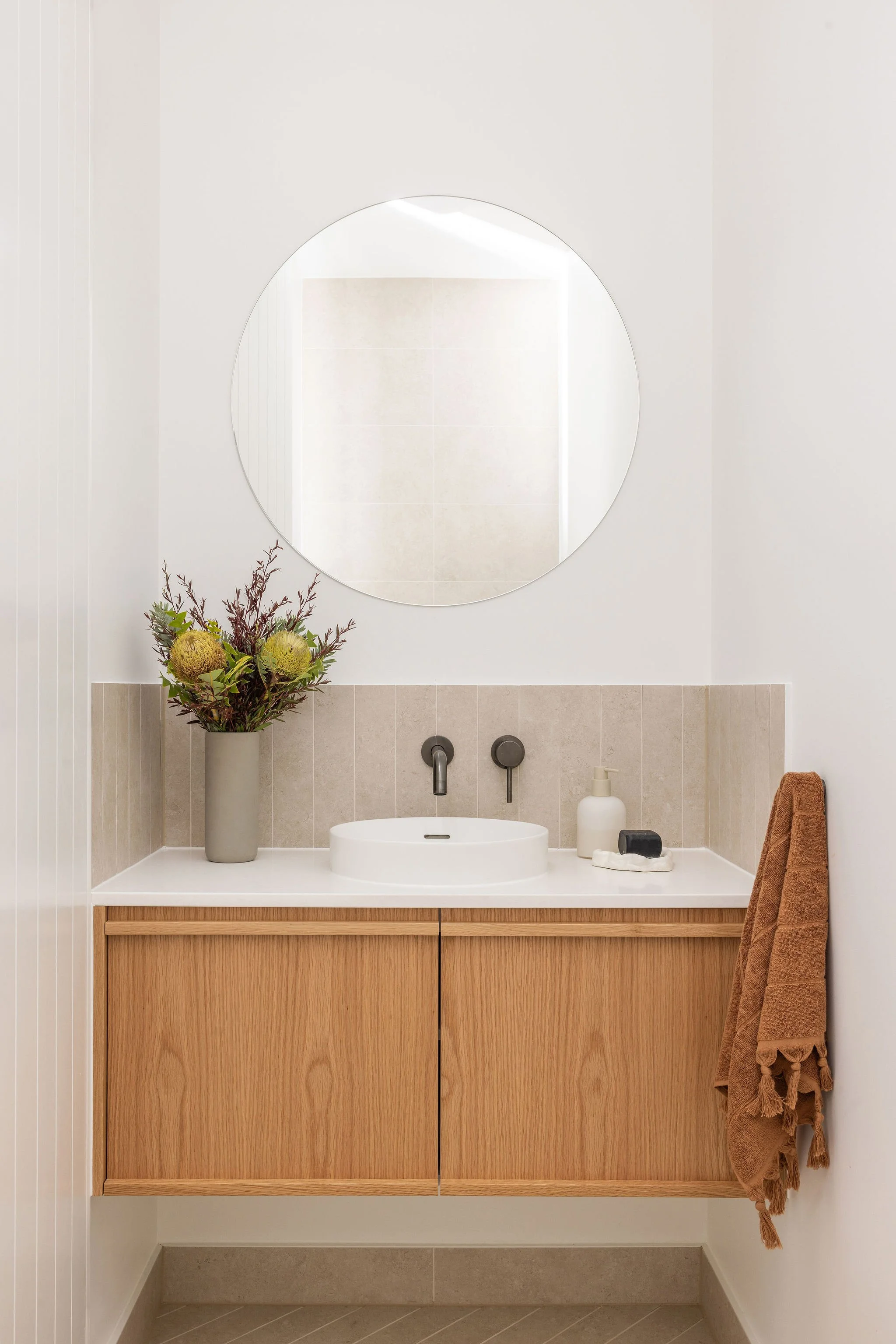 Bathroom with a round mirror above a sink, a beige vanity, and a tall white vase with green and brown flowers. There are soap and a towel on the countertop, and a brown towel hanging on a hook to the right.