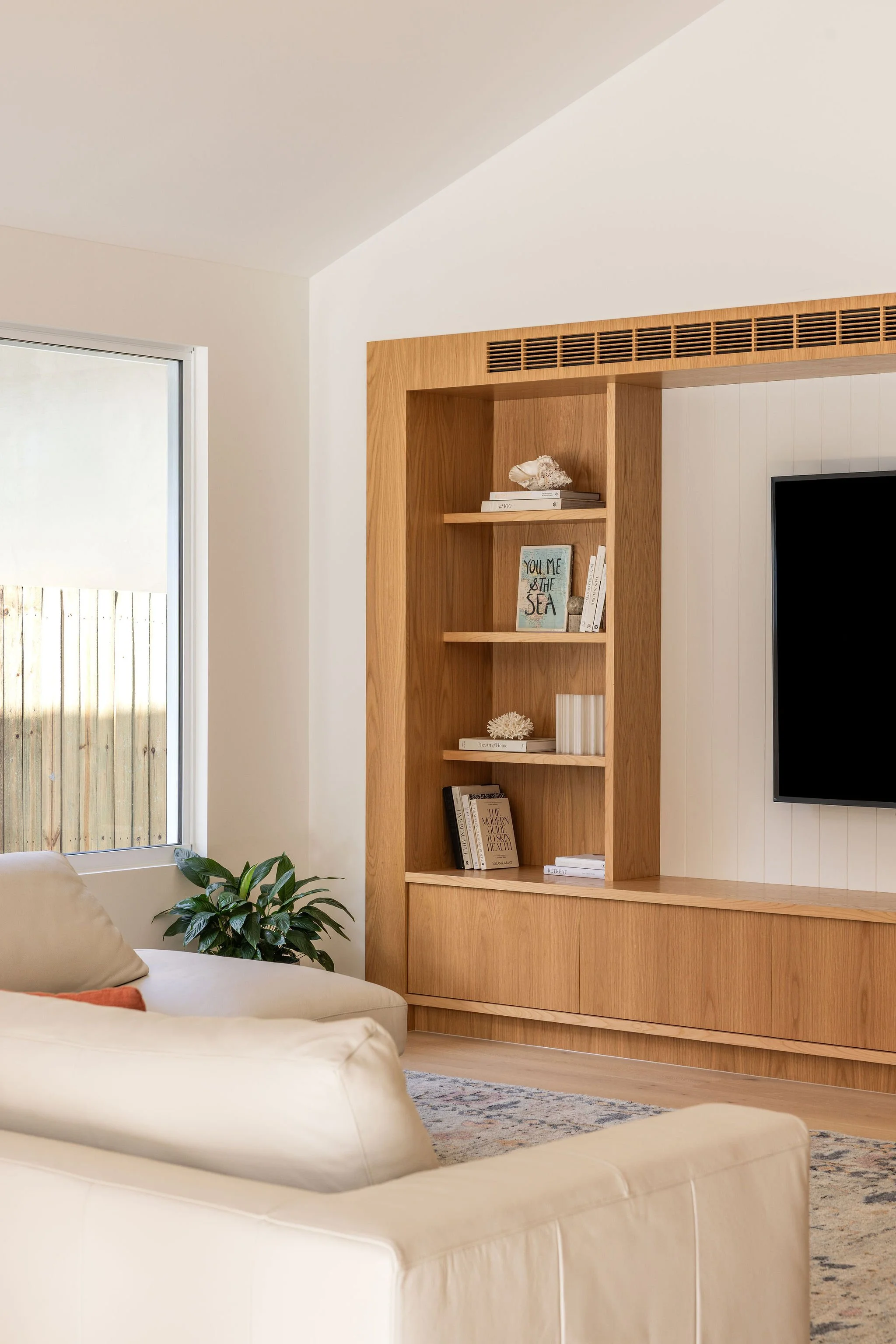 Living room with cream sofa, green plant, wooden built-in bookshelf with books and decorative items, window with wooden fence outside, patterned rug, and TV.