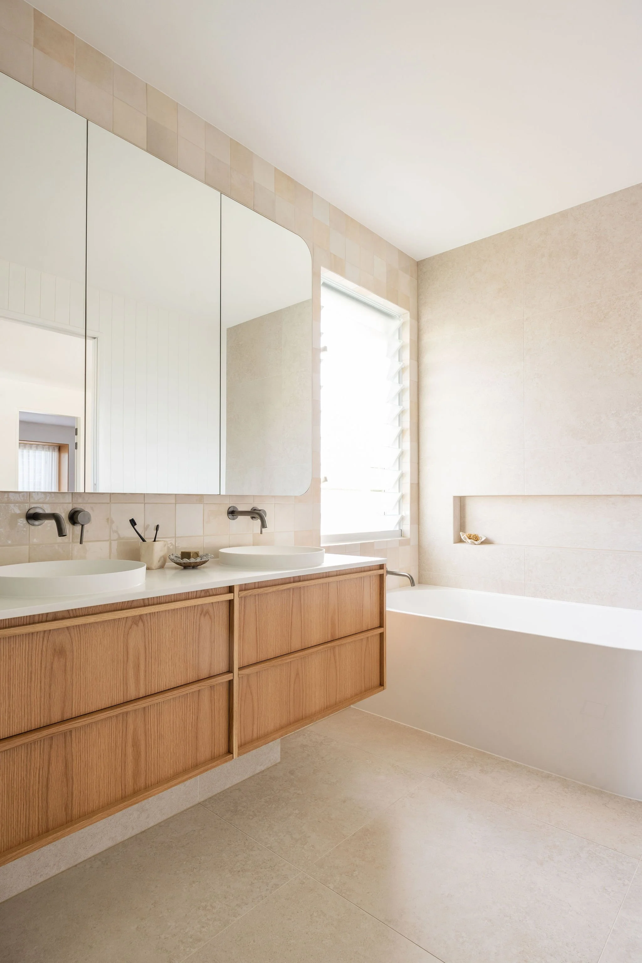 Modern bathroom with a wooden vanity, two white vessel sinks, round black faucets, large rectangular mirror, beige tiled wall, window with white blinds, and beige tiled floor.