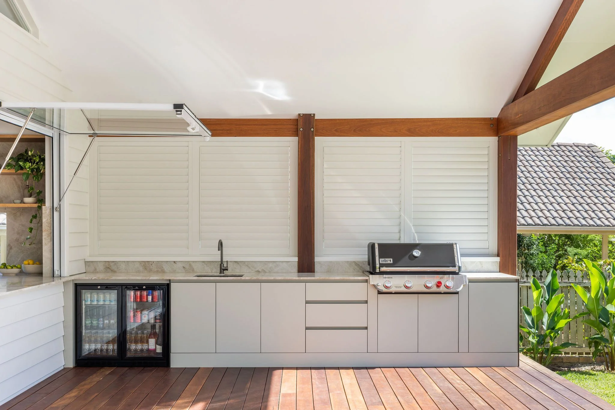 A modern outdoor kitchen with a countertop, built-in grill, small refrigerator, and cabinets, surrounded by wooden beams and a sloped roof, with lush greenery outside.