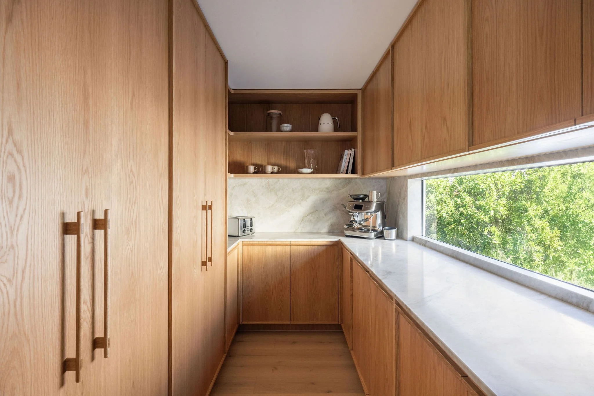 Modern kitchen with wooden cabinets, marble countertop, and large window showing greenery outside.