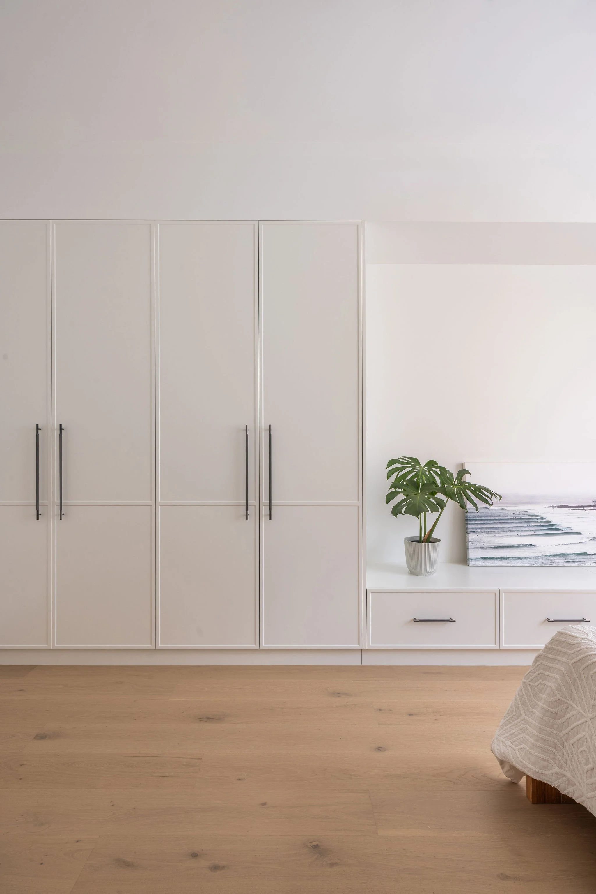 A minimalist room with white built-in cabinets, a potted plant on a small white table, and a piece of artwork featuring water and a mountain in the background. The room has a light wood floor and a neutral color palette.