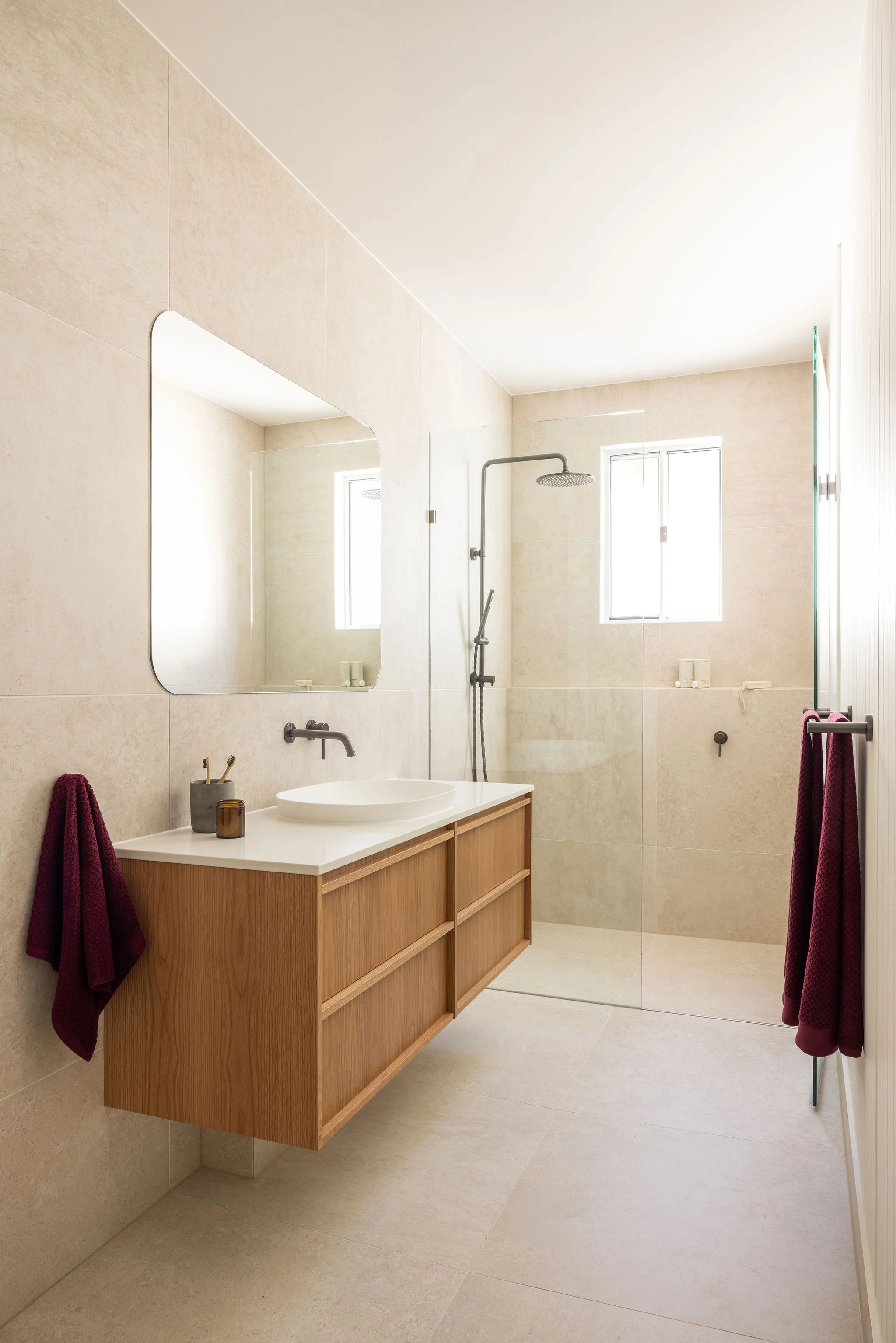 Modern bathroom with a wooden vanity, a mirror, and a walk-in shower with a rain shower head. Burgundy towels hang on hooks on either side of the vanity. Natural light comes through two windows.