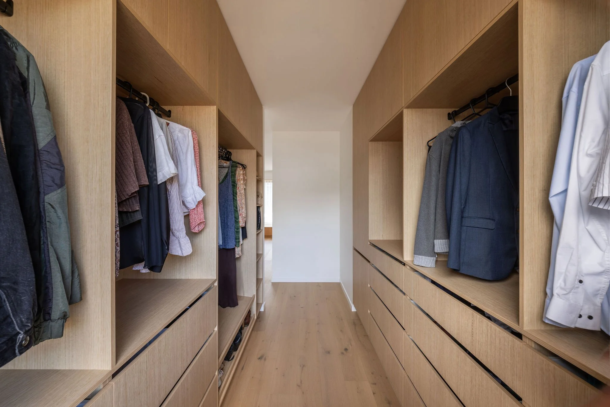 View of a walk-in closet with wooden shelves and hanging clothes, including jackets, shirts, and pants, in a modern home.