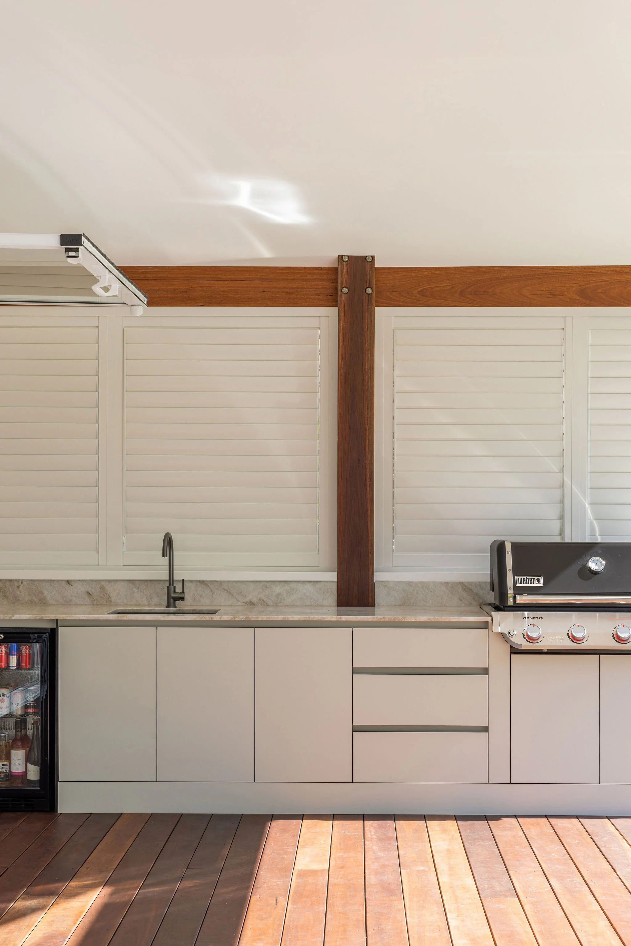 Modern outdoor kitchen with a wooden beam ceiling, white cabinet doors, a built-in grill, a sink, and a small refrigerator, all on a wooden deck.