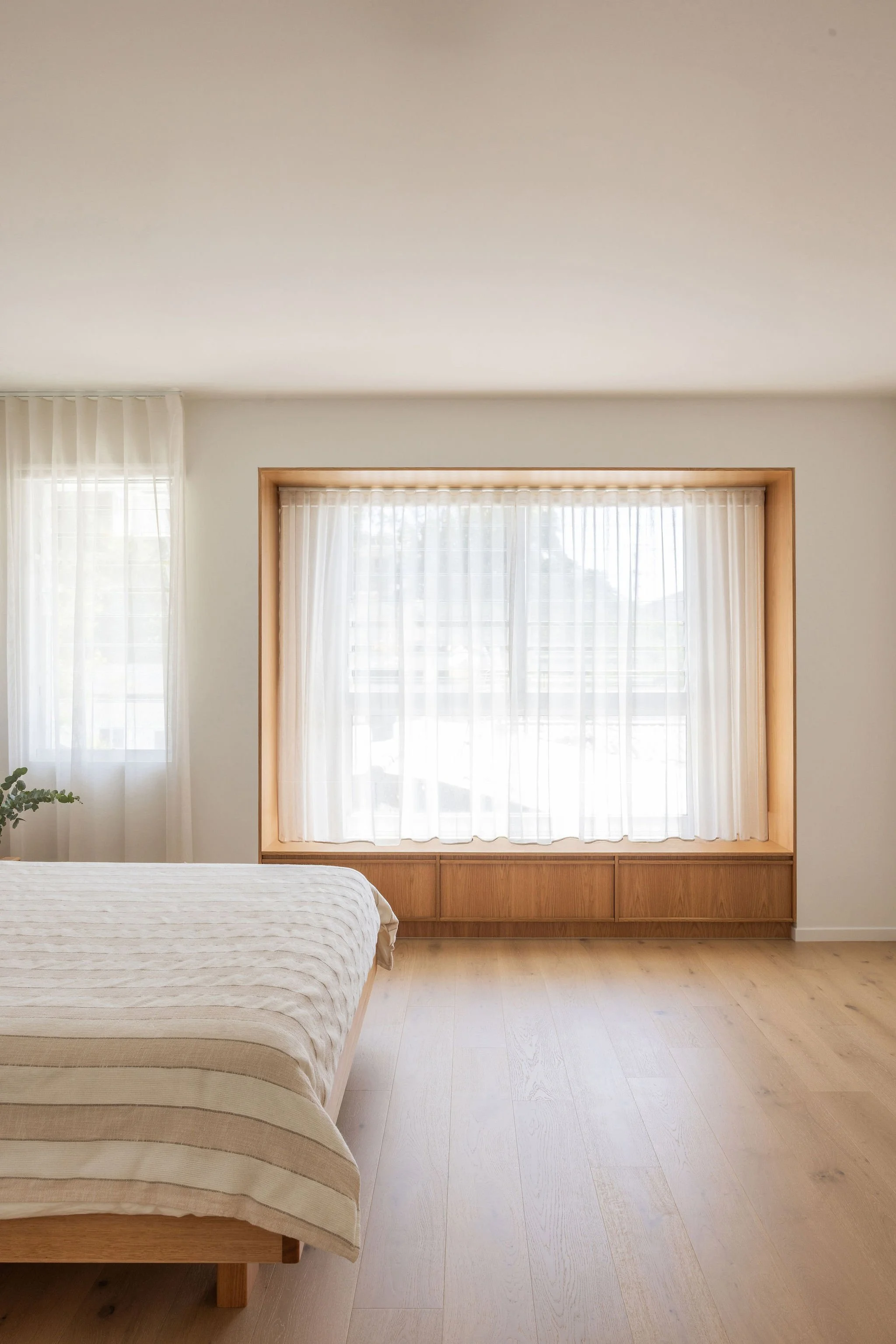 A bedroom with a bed and light-colored wooden floor, large window with sheer curtains, and a wooden frame around another window or nook.