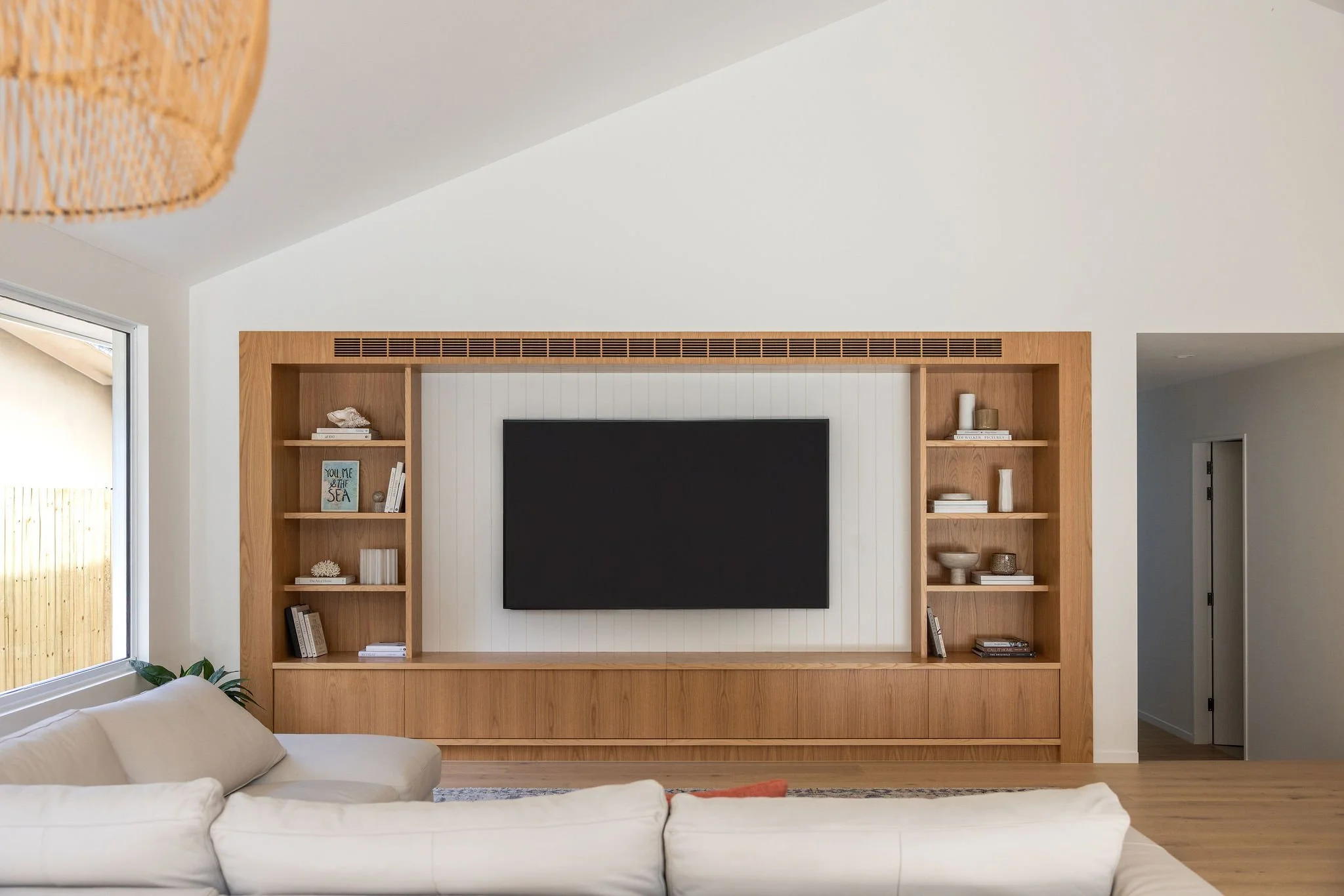 Living room with a beige couch, a large wooden entertainment center with shelves holding books and decorative items, a flat-screen TV mounted on wall, and a large window with a wooden fence outside.