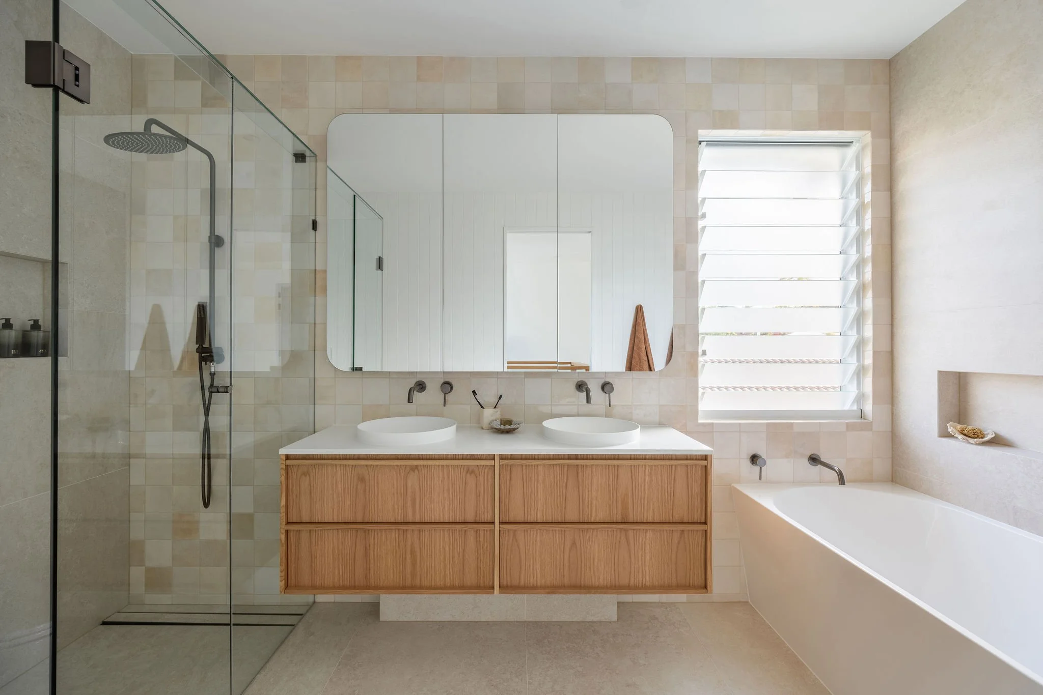 Modern bathroom with a double vanity, white vessel sinks, wood cabinet, large mirror, walk-in shower with glass enclosure, window with white louvers, bathtub, and neutral beige tiles.