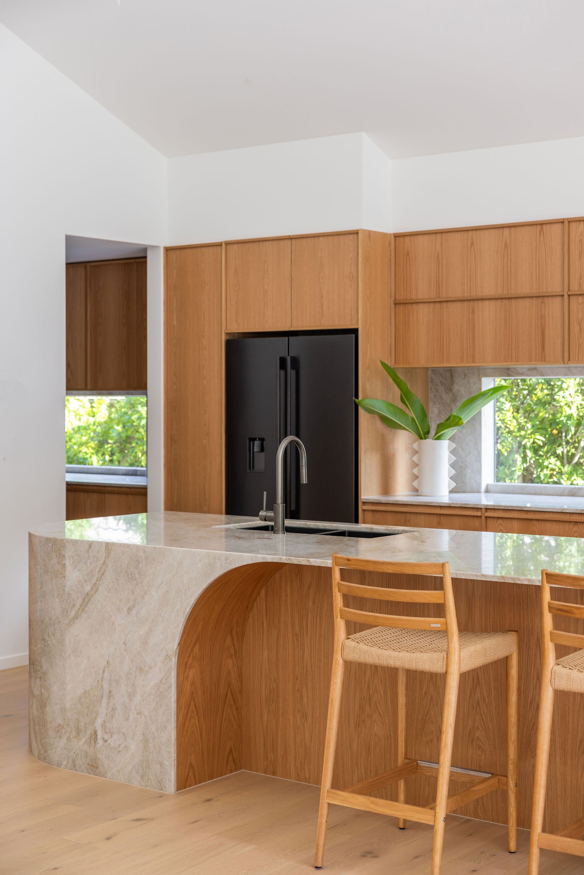 Modern kitchen with wooden cabinetry, a black refrigerator, a marble kitchen island with a sink, a white vase with greenery, and windows showing outdoor greenery.