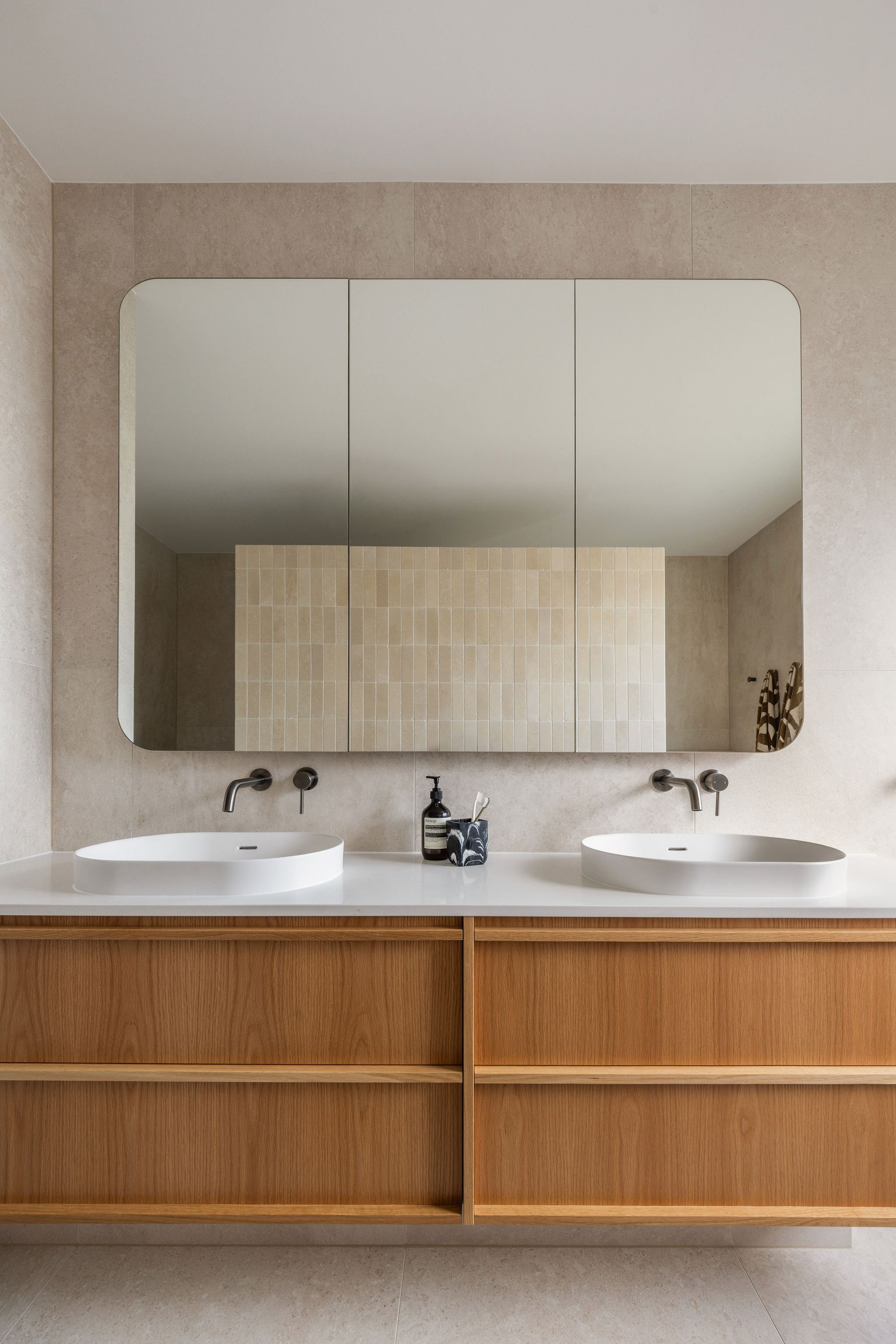 Modern bathroom with a double vanity, two oval sinks, wooden cabinet, large mirror, beige tiled walls, and small toiletries on the counter.