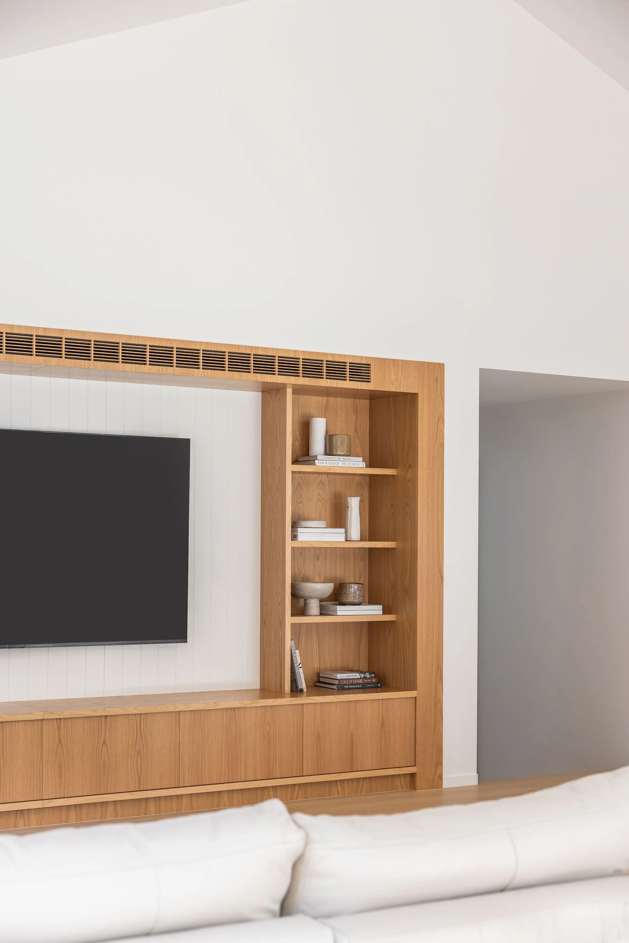 Interior view of a modern living room with a mounted black flat-screen TV on a white paneled wall. To the right of the TV is a wooden built-in shelving unit with decorative vases, books, and bowls. The ceiling is sloped, and there's a gray wall visib