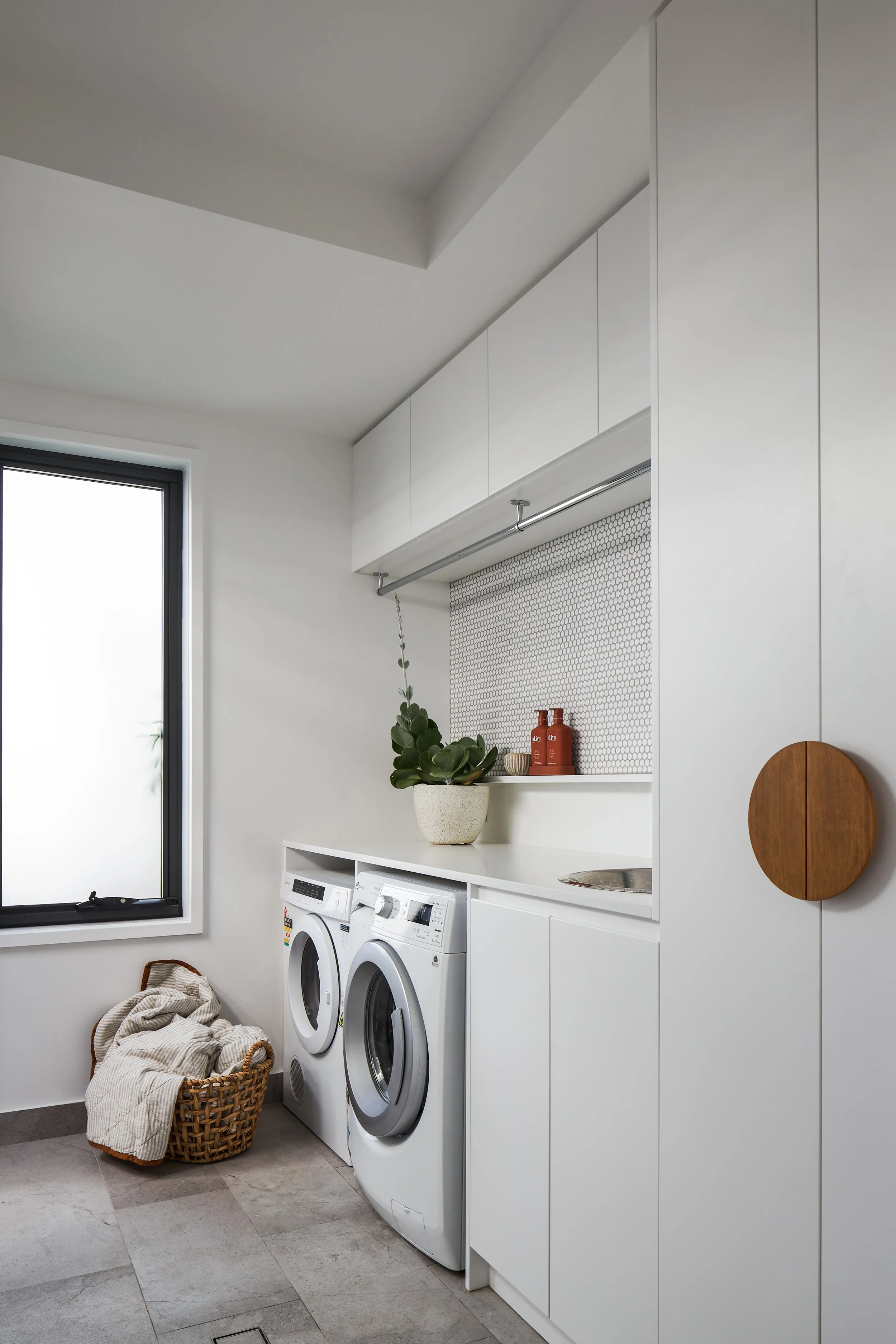 A laundry room with a window, white cabinets, a washer and dryer, a basket of laundry, a plant, and some decorative bottles on a shelf.