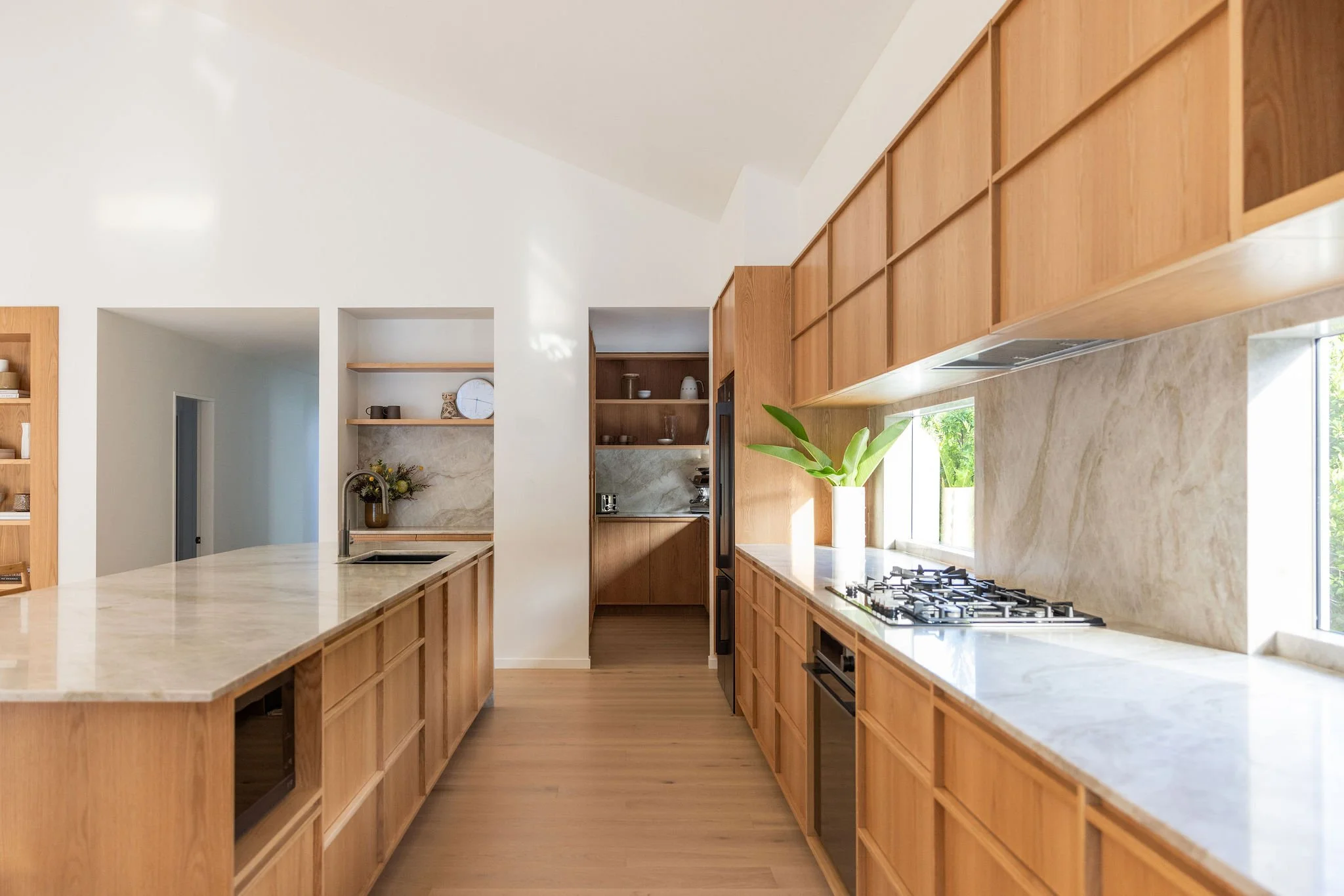 A modern kitchen with wooden cabinets, a marble countertop, and a gas stove. There is a small potted plant on the counter and open shelves with dishes. Large windows allow natural light into the space.