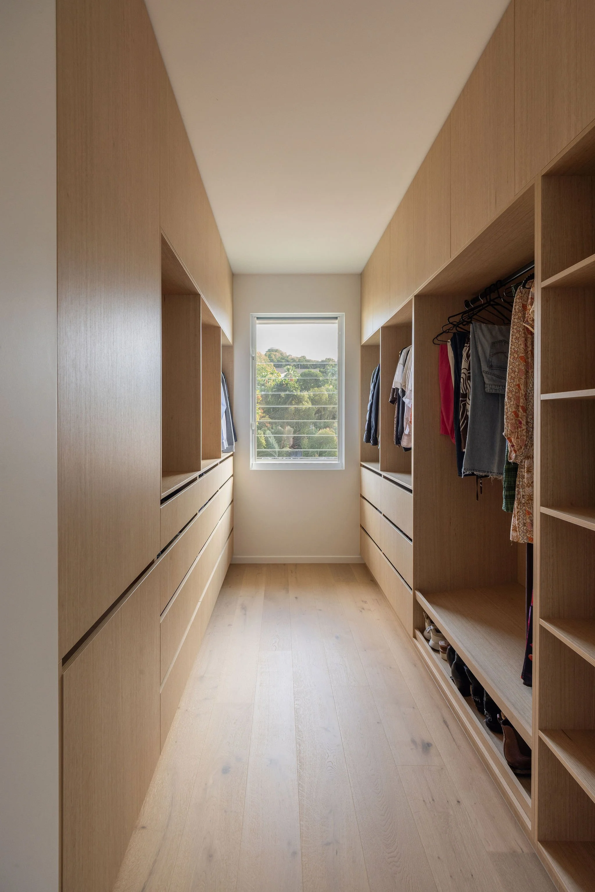 A walk-in closet with wooden cabinetry on both sides, a window at the end showing trees and a hill, with hanging clothes and shoes on the shelves.