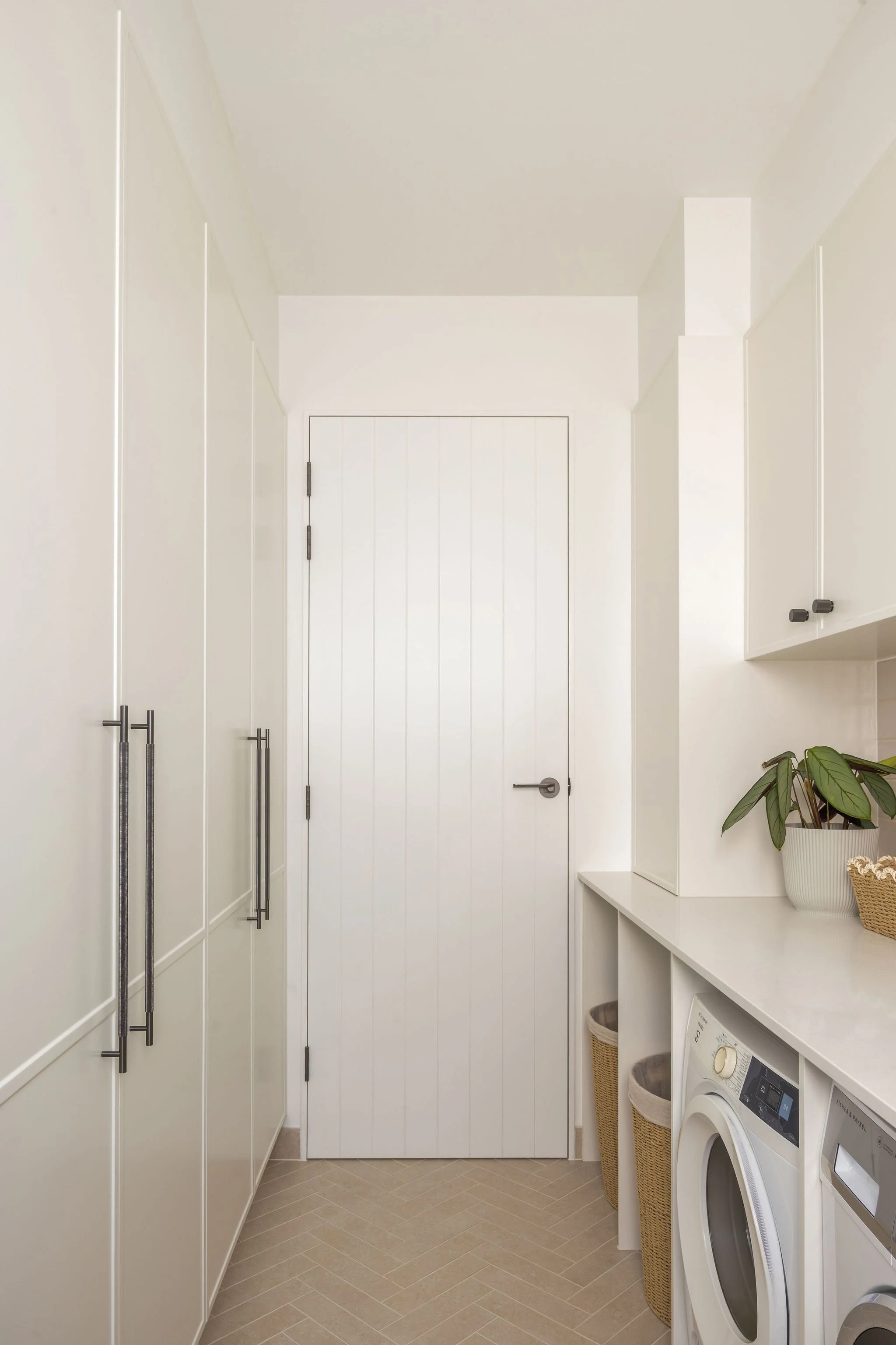 A minimalist laundry room with white cabinets and a white washing machine. There is a potted plant on a white shelf and two wicker baskets beside the washer. The floor is covered with beige tiles arranged in a herringbone pattern.