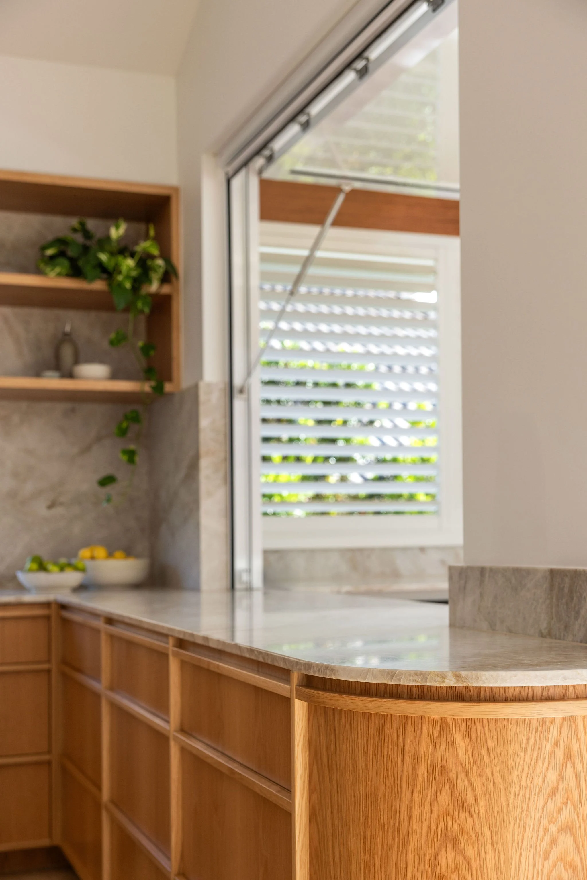 A kitchen with wooden cabinetry and a cream-colored marble countertop. There are shelves with plants and decor, and an open window with slatted shutters letting in natural light.