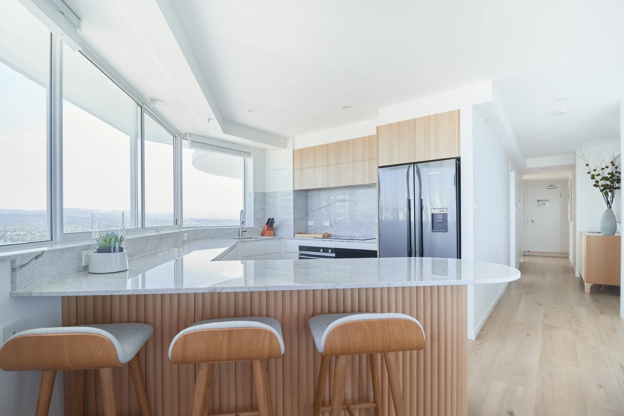 Modern kitchen with a white marble countertop, wooden cabinets, stainless steel refrigerator, and large windows offering city views.