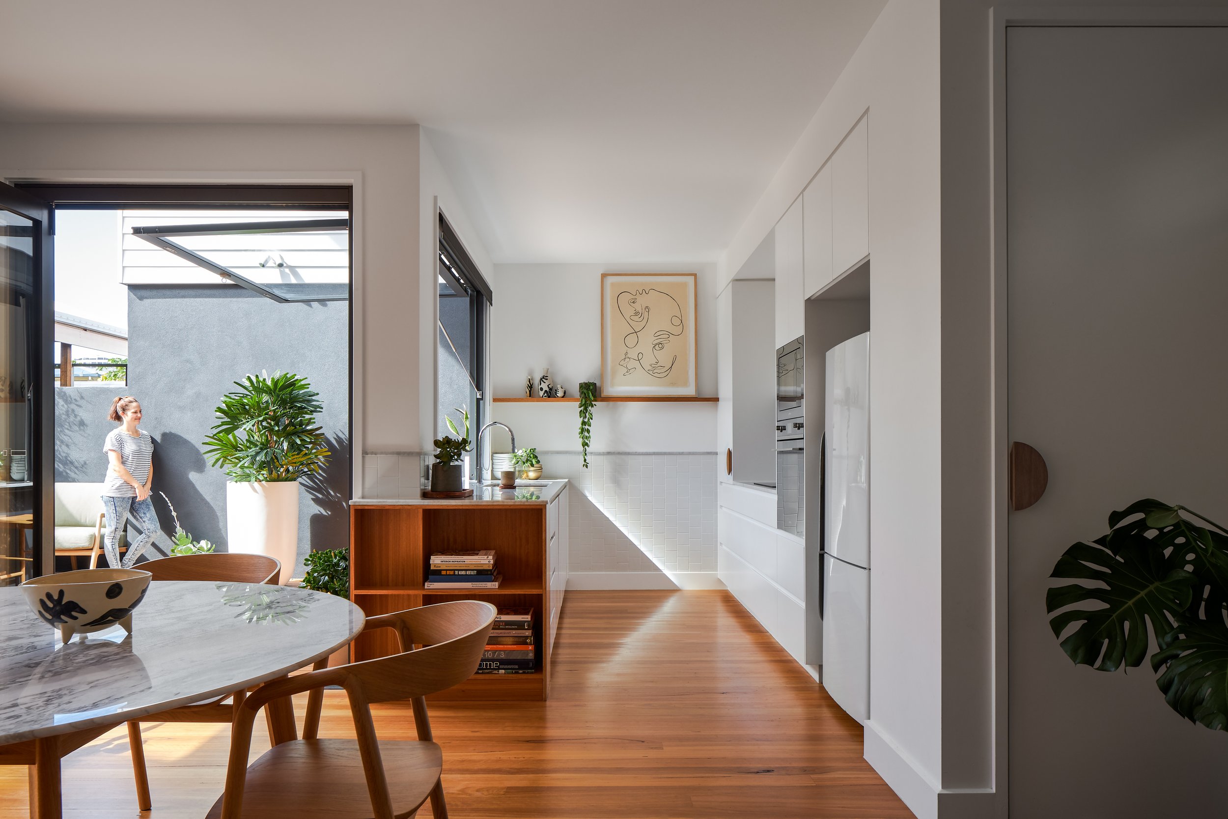 Modern open-concept kitchen and dining area with white cabinetry, wooden flooring, a marble-topped dining table with wooden chairs, a large potted plant outside the open door, and artwork on the wall.
