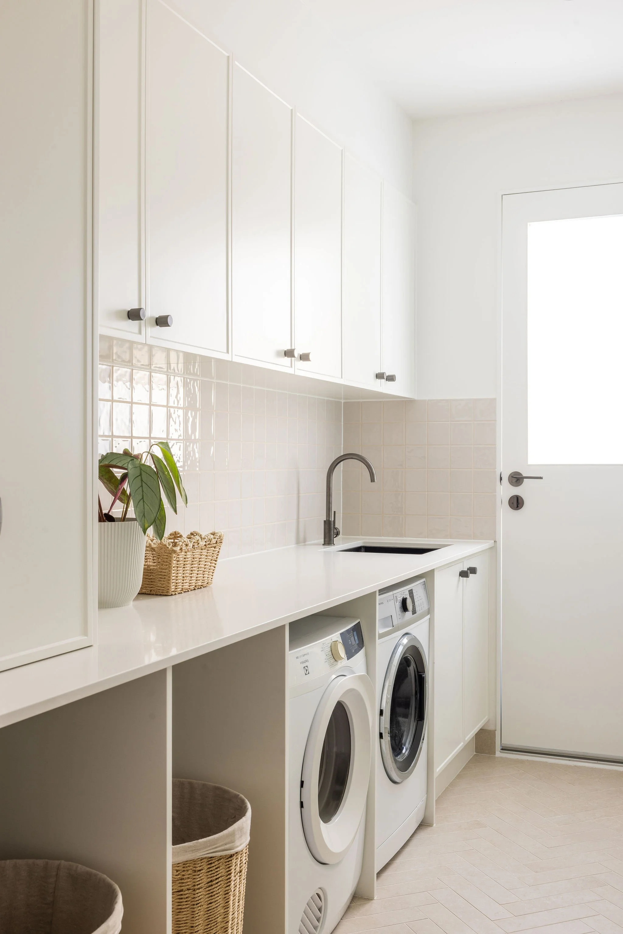 A bright white laundry room with cabinets, a countertop, a plant, a laundry basket, and two front-loading washing machines. The room has a large window and a door.